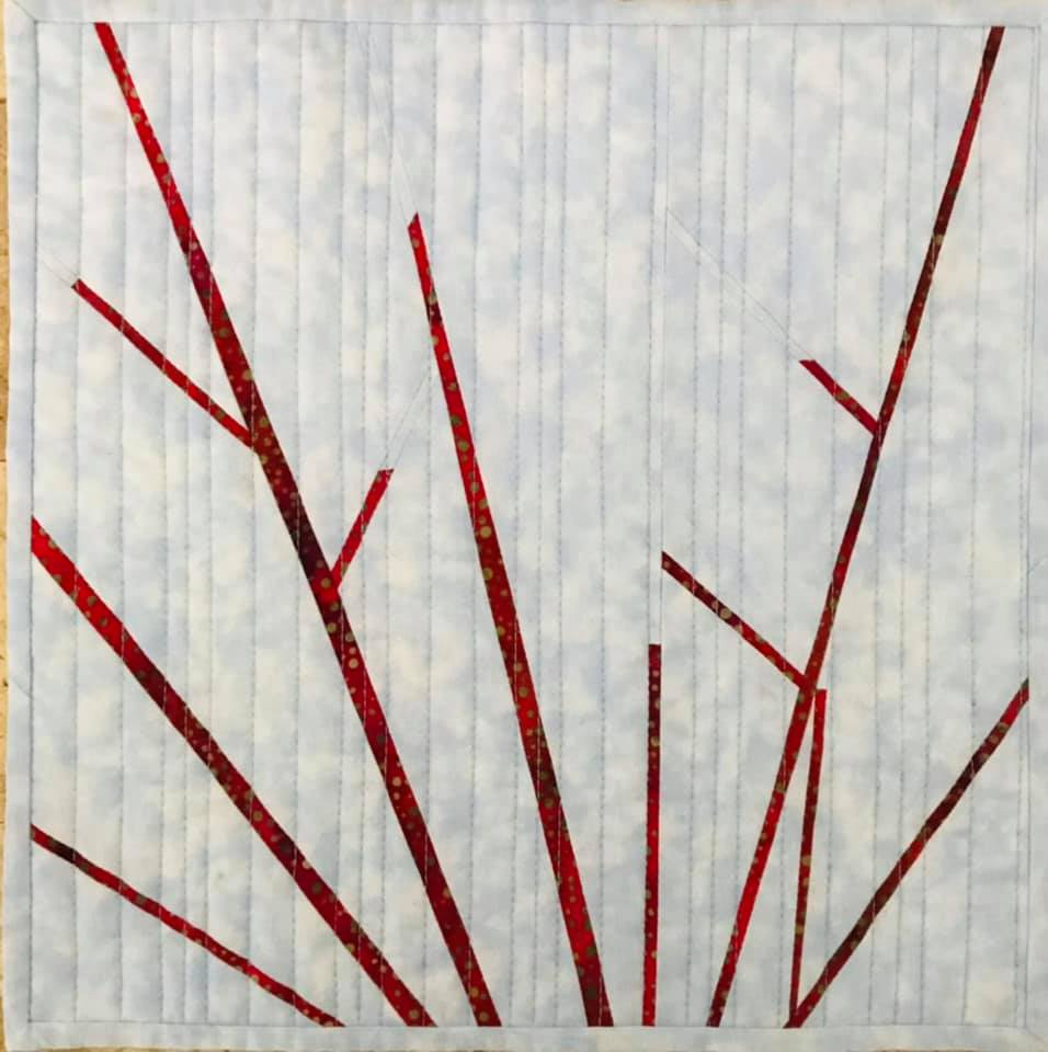 A small, square quilt with a light grey background and a bold, abstract design. The design features red lines radiating outward from the centre, resembling branches or a firework explosion.