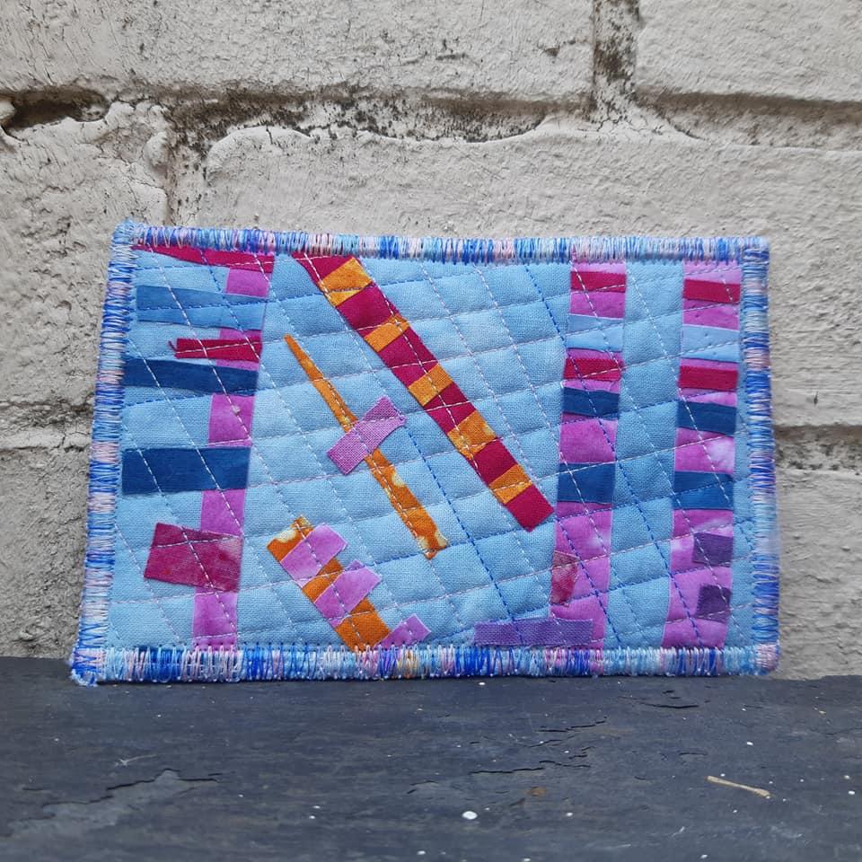 A small, rectangular quilt with a patchwork design. It features a blue background with strips of pink, purple, and orange fabric arranged in a random pattern. The quilt has a colourful, stitched border.