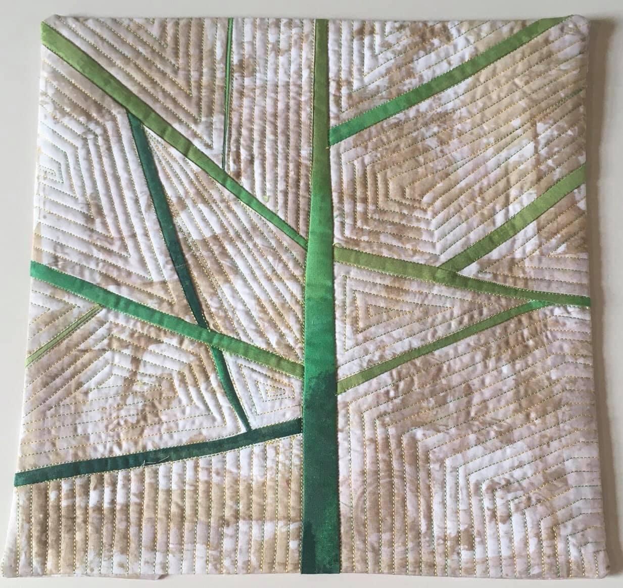 A small, square quilt with a tan background and a green tree-like design. The tree has a thick green trunk and branches that extend outward. The background is filled with a quilted pattern of small triangles.