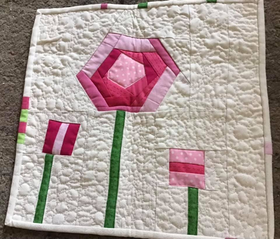A small, square quilt with a white background and a floral design. There are three flowers, one large pink flower in the center and two smaller pink and white flowers on either side. The flowers have green stems. The background is quilted with a small, white pattern.