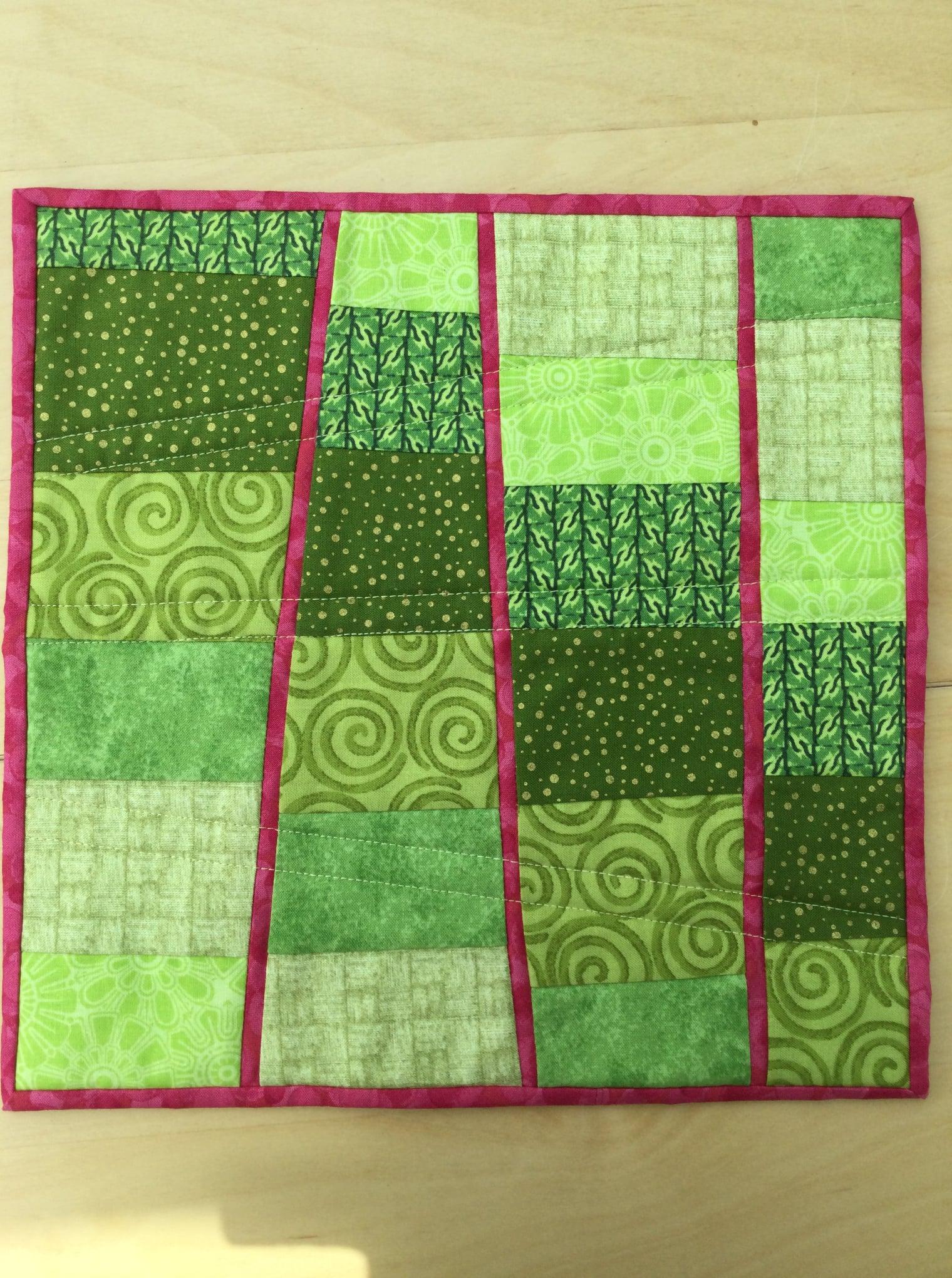 A small, rectangular quilt with a patchwork design. It features various shades of green fabric arranged in vertical strips, separated by curved seams. The quilt has a pink border.