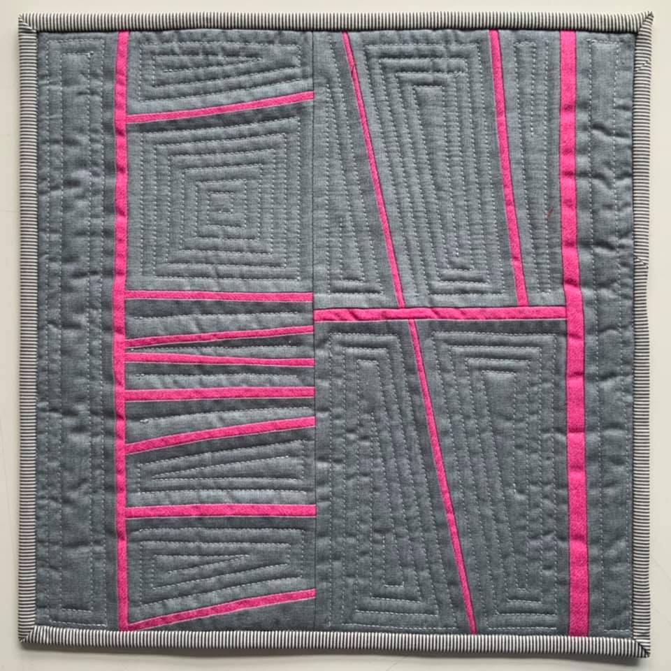 A small, square quilt with a grey background and a bold, abstract design of pink lines. The lines create various shapes and patterns, including rectangles and spirals. The quilt is framed by a thin, striped border.