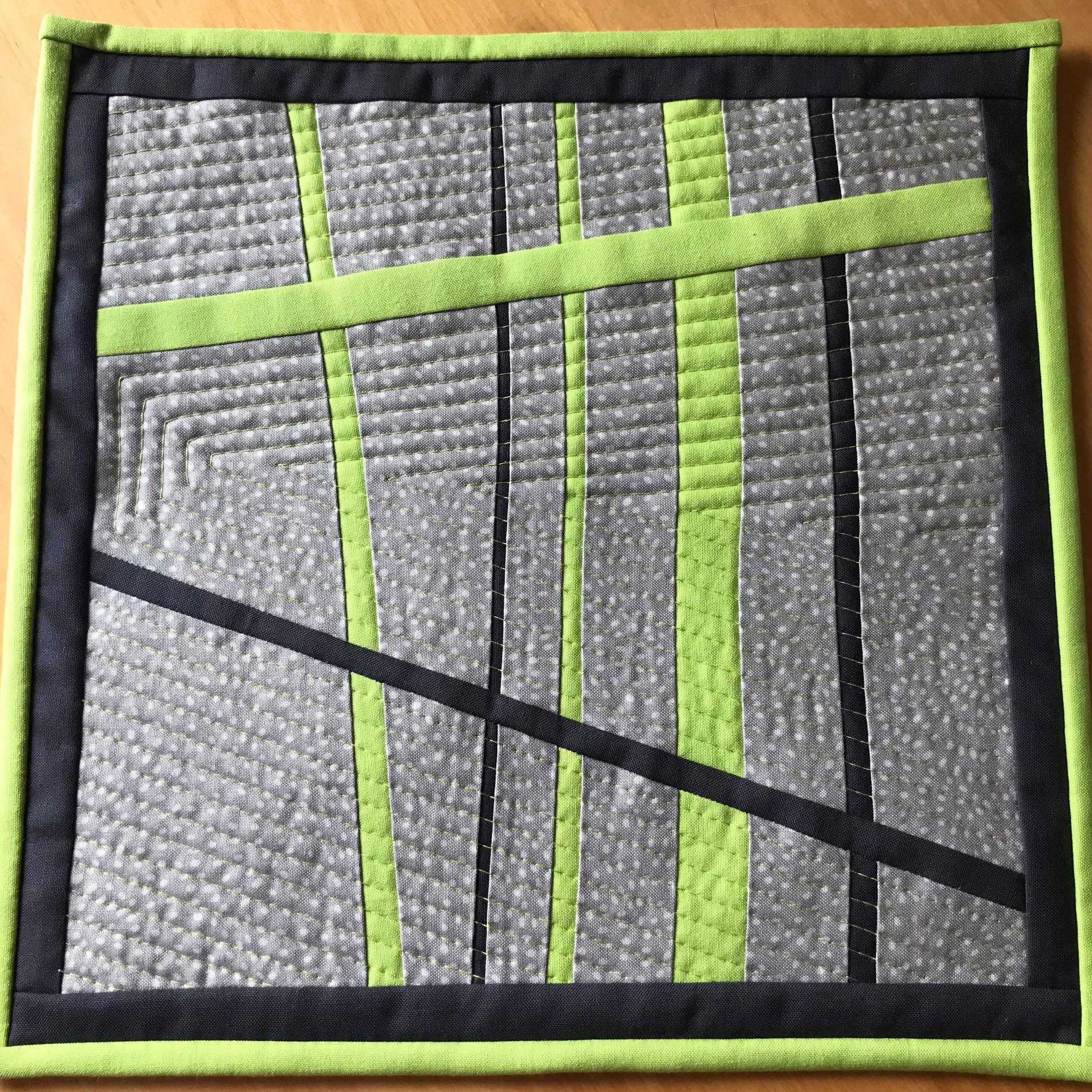 A small, square quilt with a grey background and a green border. The quilt features a geometric design with intersecting black and green lines, creating a pattern of rectangles and triangles. The background fabric has a subtle textured pattern.