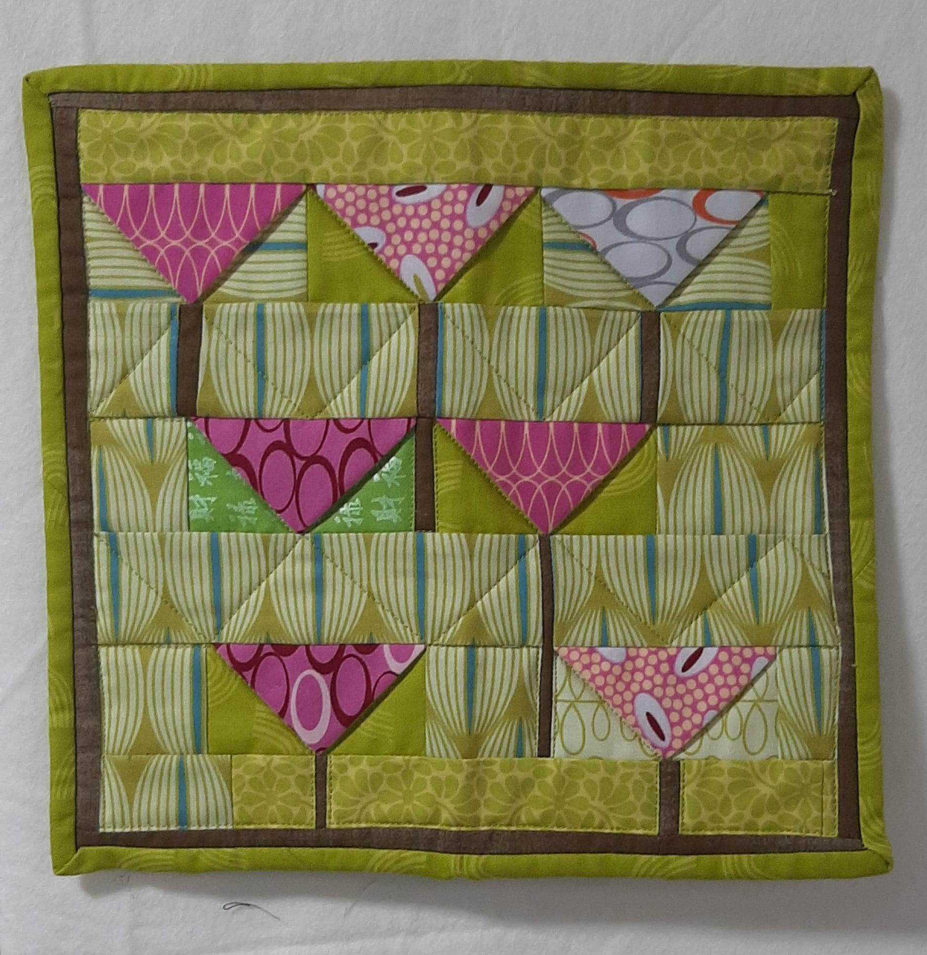 A small, rectangular quilt with a patchwork design. It features a green background with pink and white shapes arranged in a pattern resembling flowers. The quilt has a brown border.