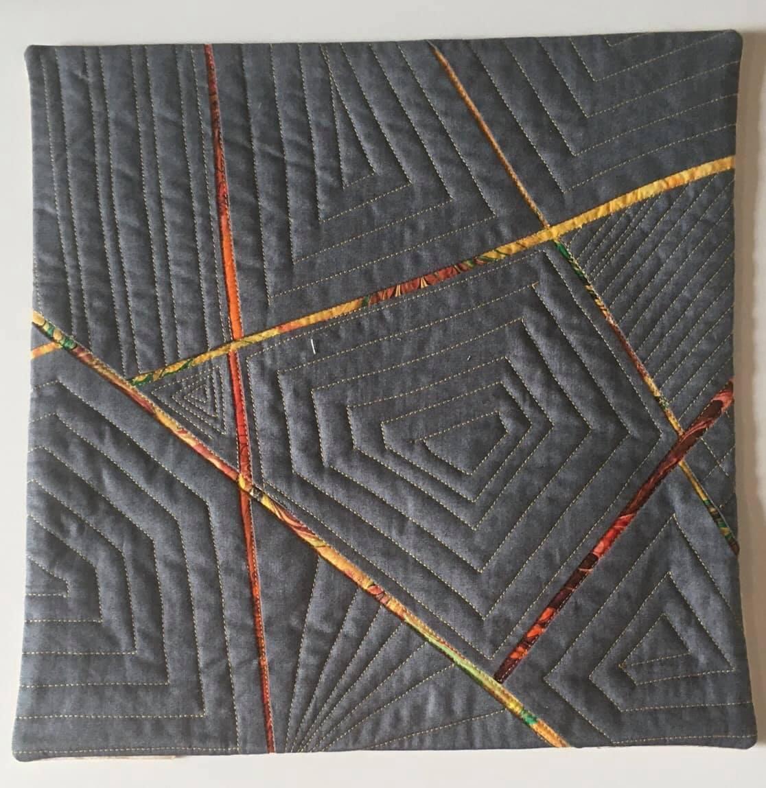 A small, rectangular quilt with a grey background. The quilt features a geometric design with intersecting lines of orange and yellow fabric. There are also smaller, nested shapes within the larger design, creating a complex pattern.