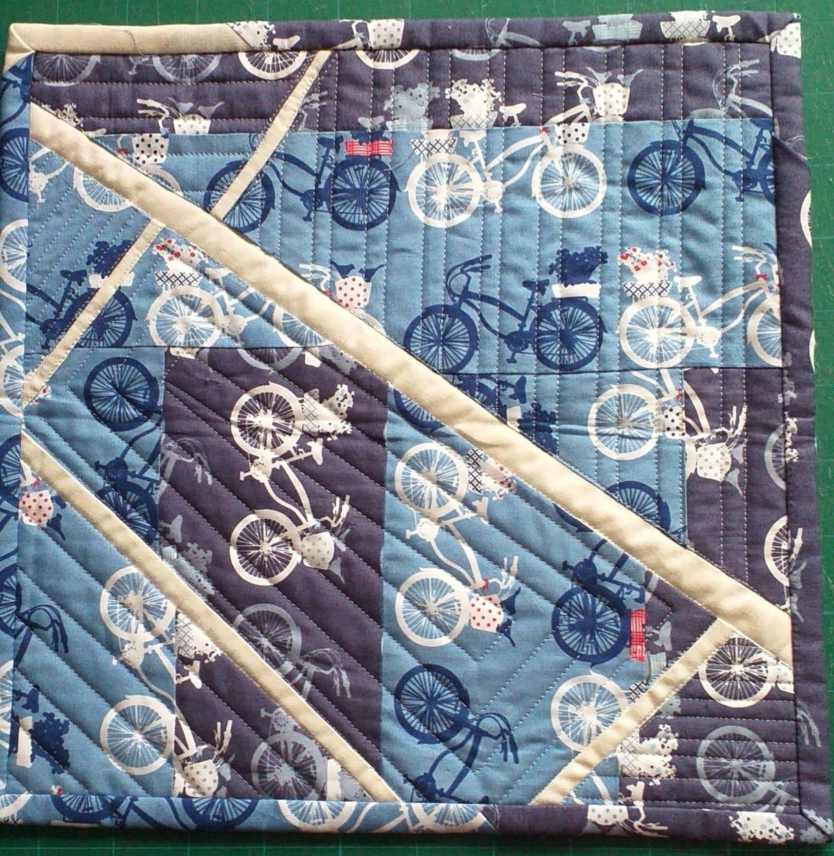 A small, square quilt with a patchwork design. It features a blue fabric with a pattern of bicycles, cut into geometric shapes and arranged in a diagonal pattern. The quilt has a white border and a dark blue backing.