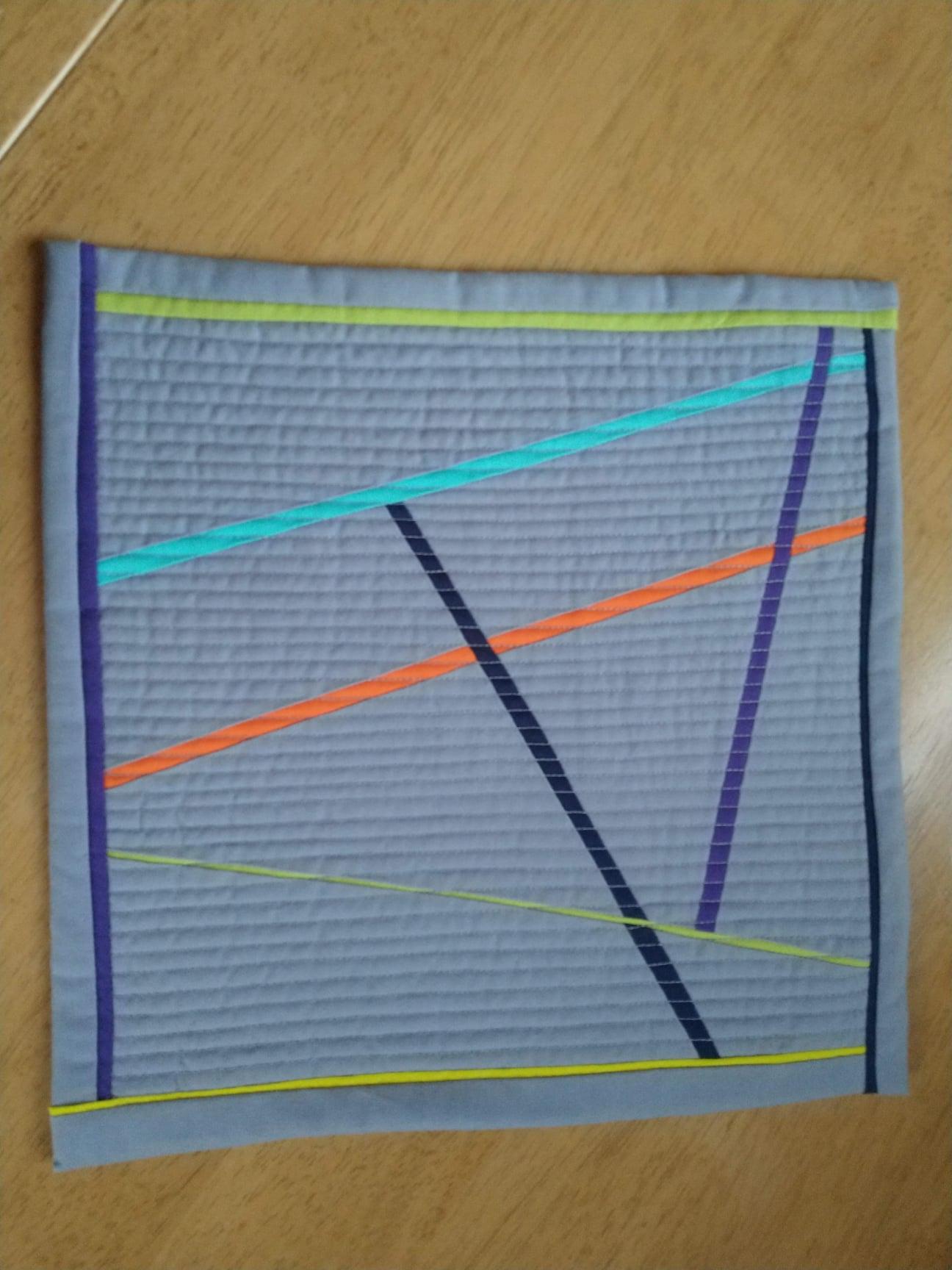 A small, rectangular quilt with a grey background and a colourful geometric design. The design features intersecting lines in orange, blue, purple, and black. The quilt has a green border.