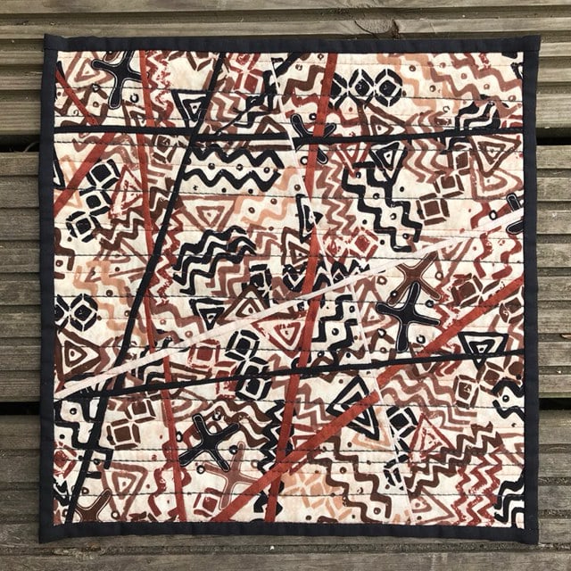 A small, square quilt with a patchwork design. It features a combination of brown, black, and white fabrics with geometric patterns and symbols. The quilt has a black border.