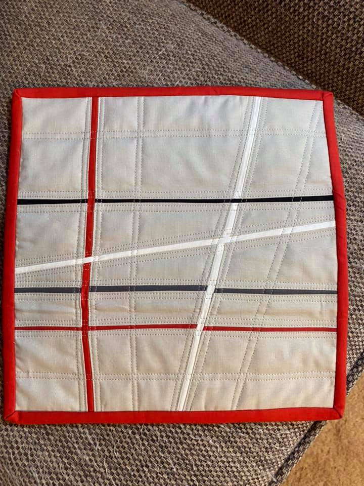 A small, rectangular quilt with a light gray background and a red border. The quilt features a geometric design with intersecting lines of red, black, and white fabric. The lines create a grid-like pattern.