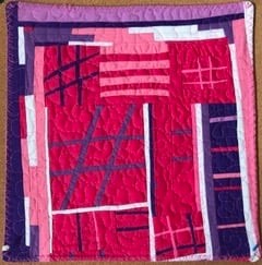 A small, square quilt with a vibrant, abstract design. It features a mix of pink, purple, and white fabrics arranged in geometric shapes and patterns. Some areas have a quilted texture, adding depth to the design.