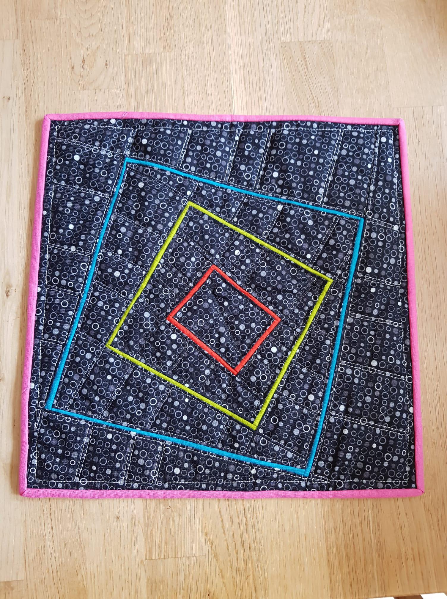 A small, square quilt with a black background and a colourful geometric design. The design features a series of overlapping squares in pink, blue, yellow, and red, each outlined with a contrasting colour. The background fabric has a pattern of small white circles.