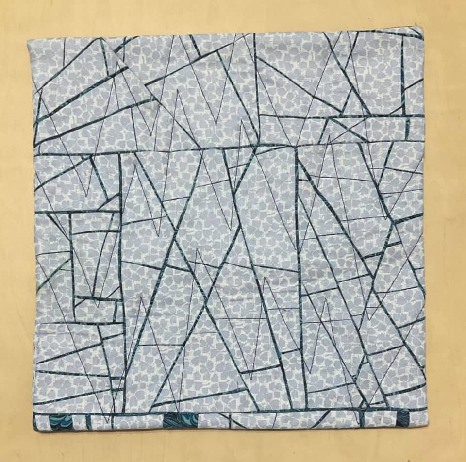 A small, square quilt with a light blue background and a dark blue border. The quilt features a geometric design with intersecting lines creating a pattern of triangles and irregular shapes. There is also a subtle, speckled pattern within the shapes.