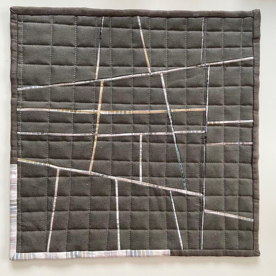 A small, square quilt with a patchwork design. The quilt is primarily dark grey with thin, intersecting lines of light grey, white, and a few pops of tan. The bottom right corner has a small strip of a light plaid fabric.