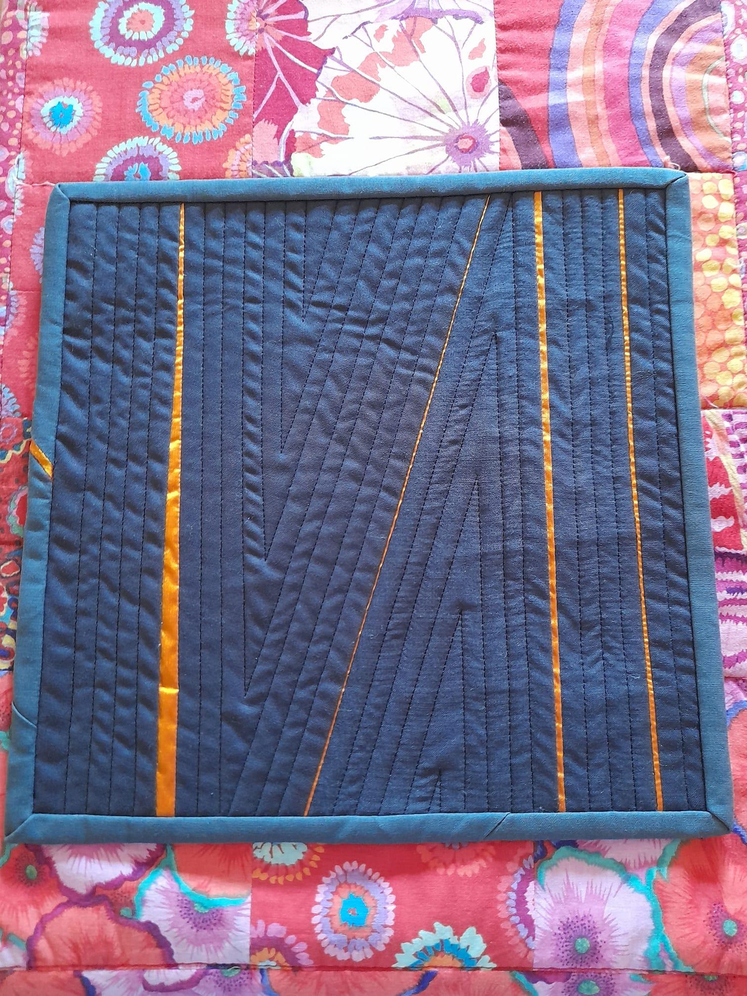 A small, rectangular quilt with a dark blue background and a teal border. The quilt features a geometric design with diagonal lines and stripes of orange fabric.