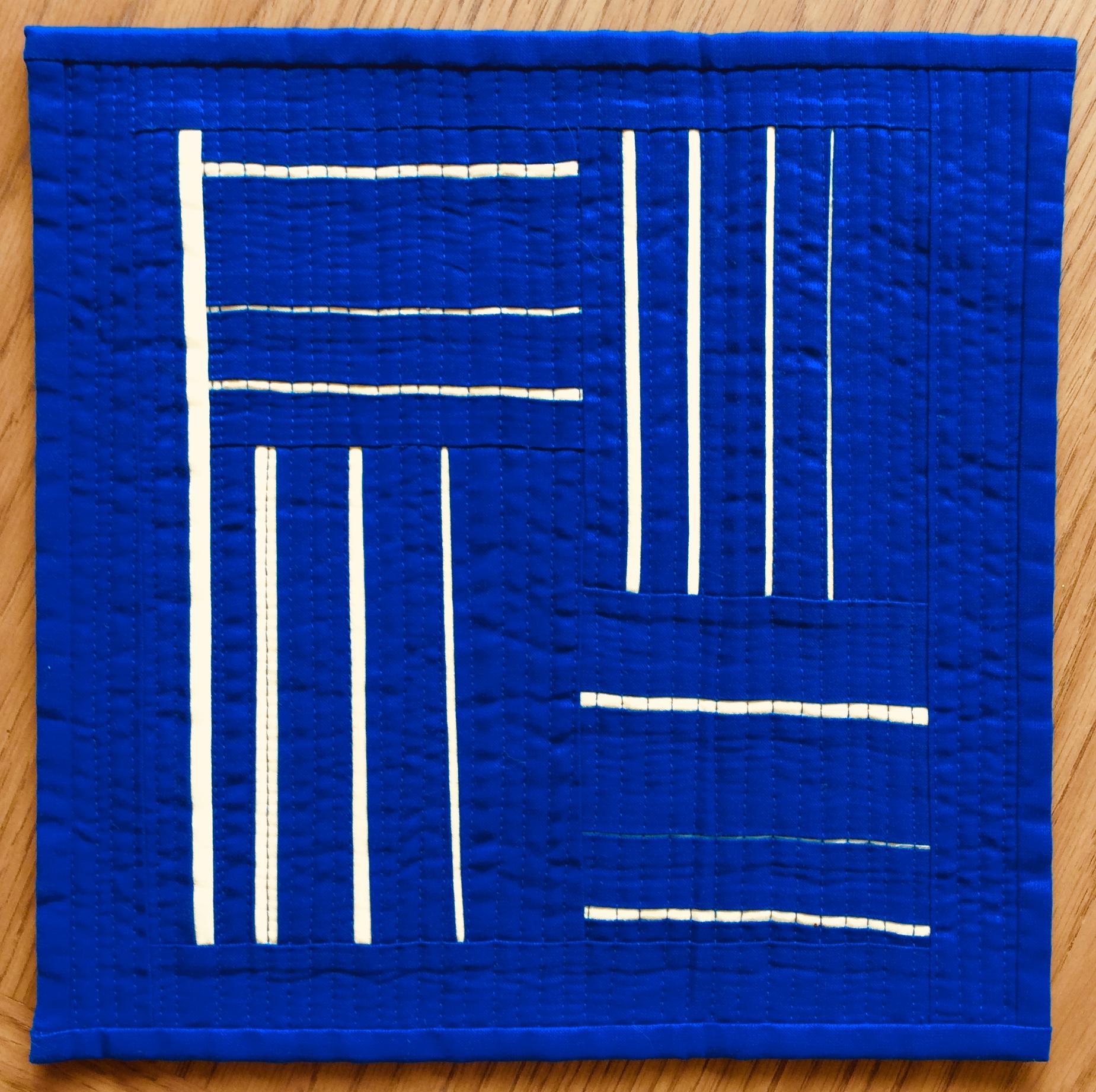 A small, rectangular quilt with a vibrant blue background. The quilt features a geometric design with white lines arranged in a grid-like pattern, creating squares and rectangles.