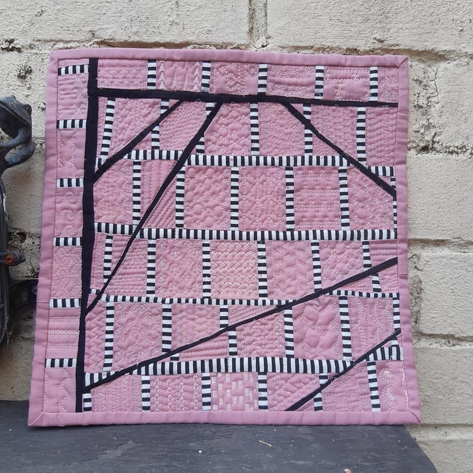A small, square quilt with a pink background. The quilt features a geometric design with black lines creating a grid-like pattern. There are also white stripes within some of the squares, adding texture to the design.