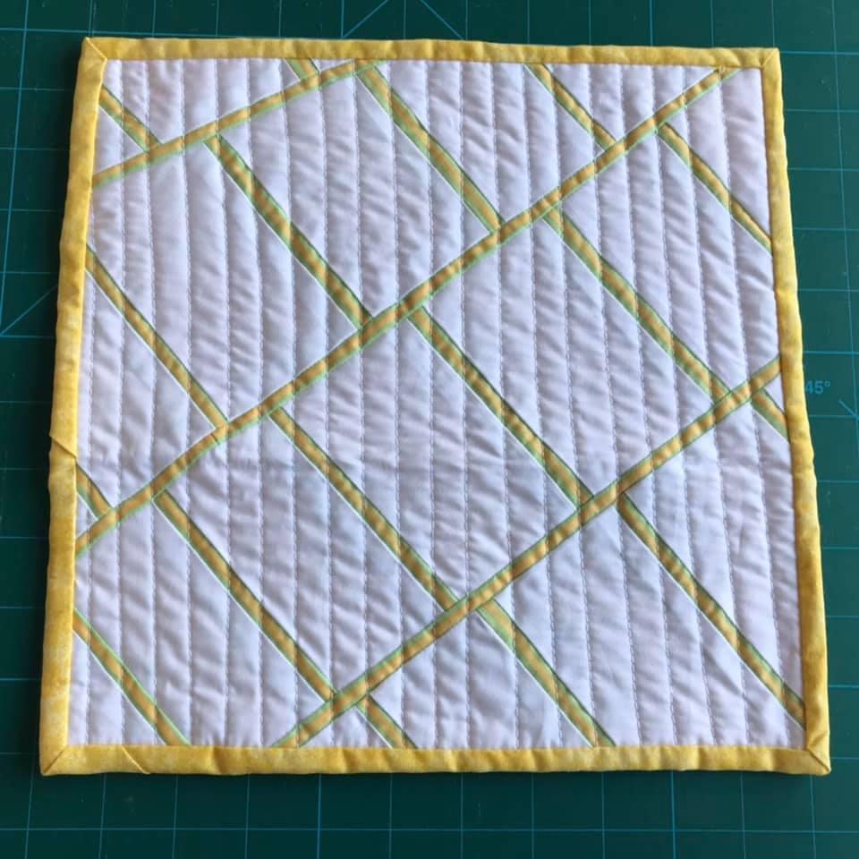 A small, rectangular quilt with a white background and a yellow border. The quilt has a geometric design with yellow lines arranged in a diamond pattern. The lines are quilted in a straight line pattern.