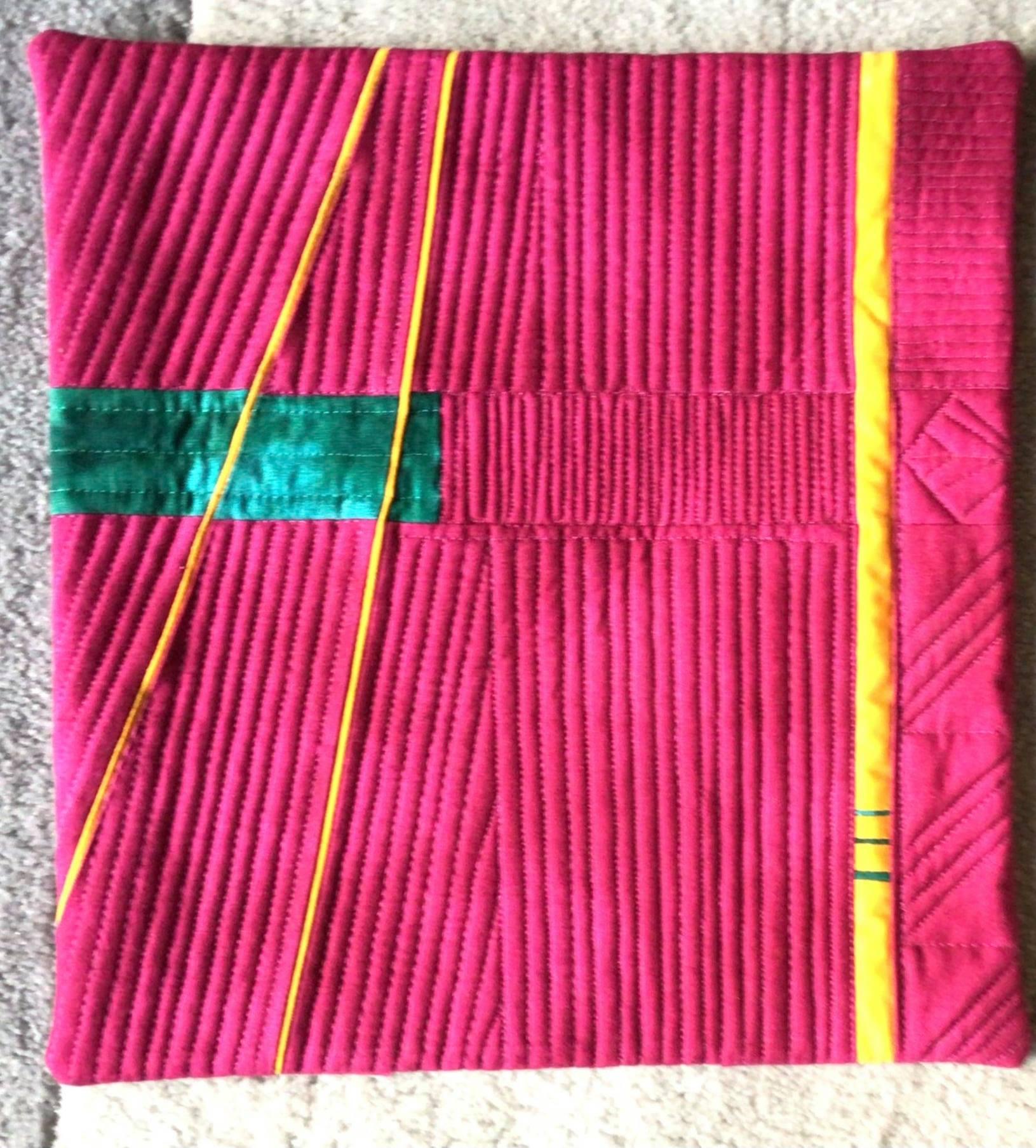 A small, rectangular quilt with a vibrant pink background. The quilt features a geometric design with yellow and green lines creating a dynamic pattern. The lines are quilted with a textured, striped pattern.
