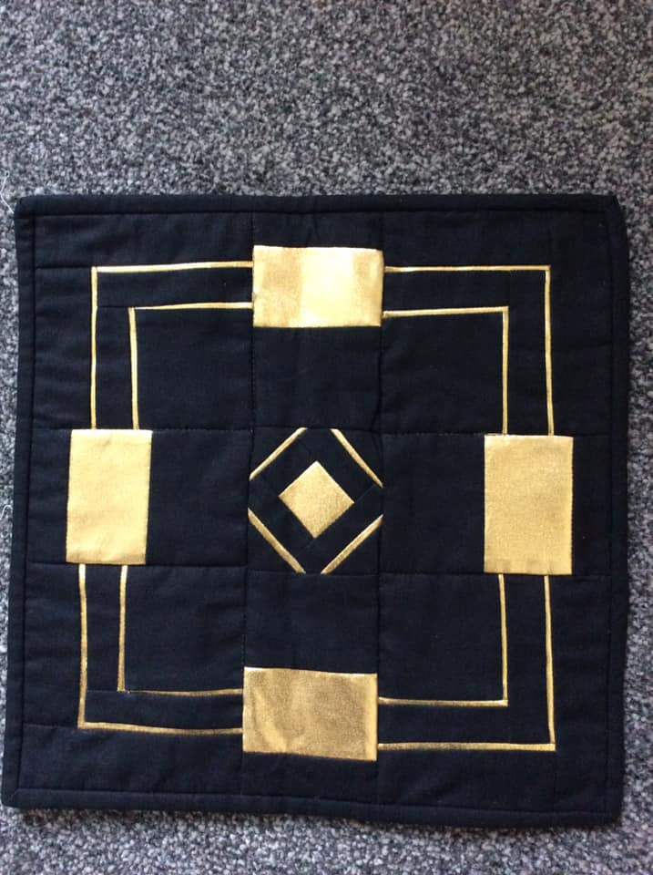 A small, square quilt with a black background and a geometric design in gold. The design features squares and diamonds arranged in a symmetrical pattern.