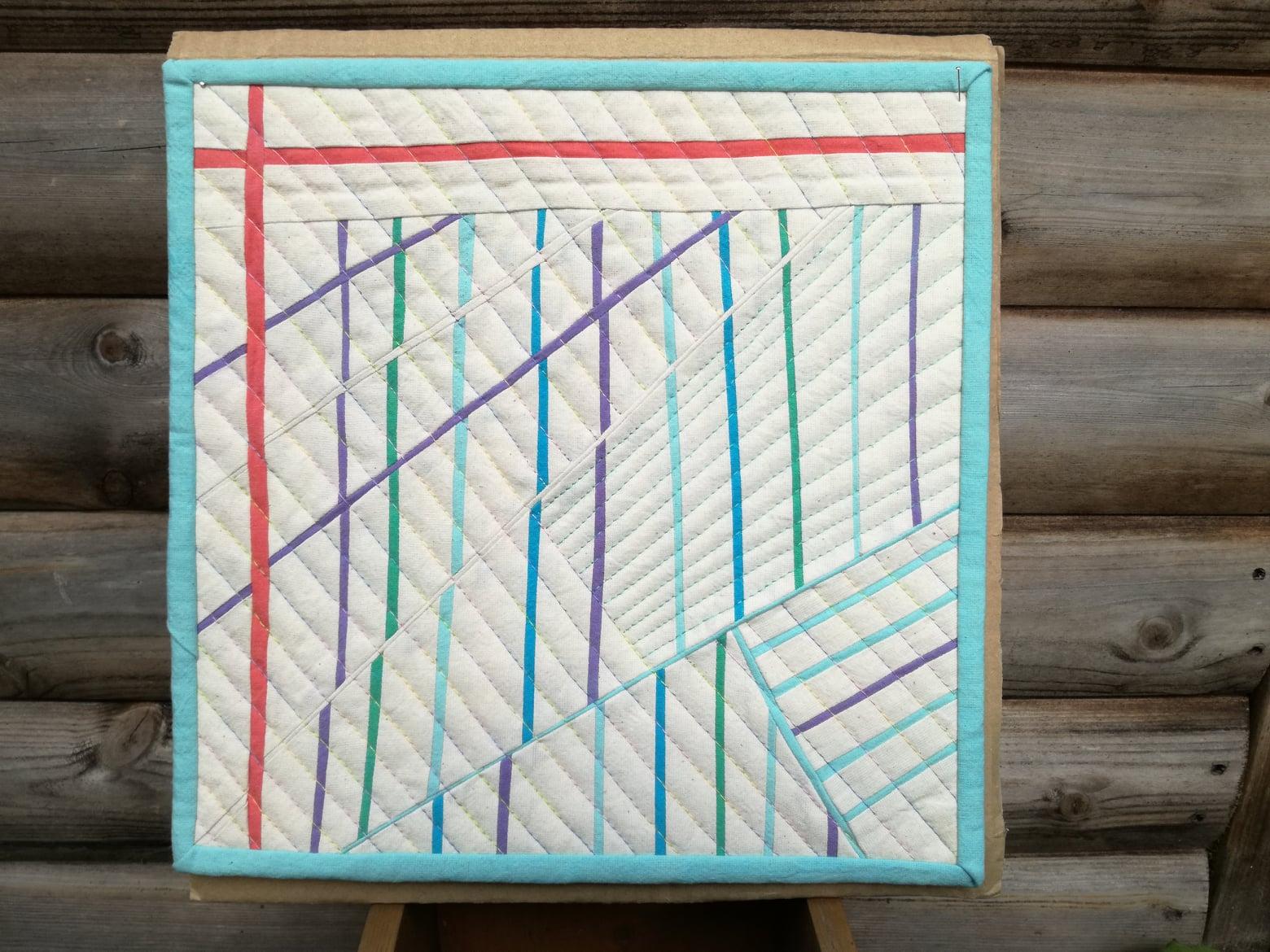 A small, square quilt with a white background and a blue border. The quilt features a geometric design with intersecting lines in various colours, including red, green, purple, and blue.