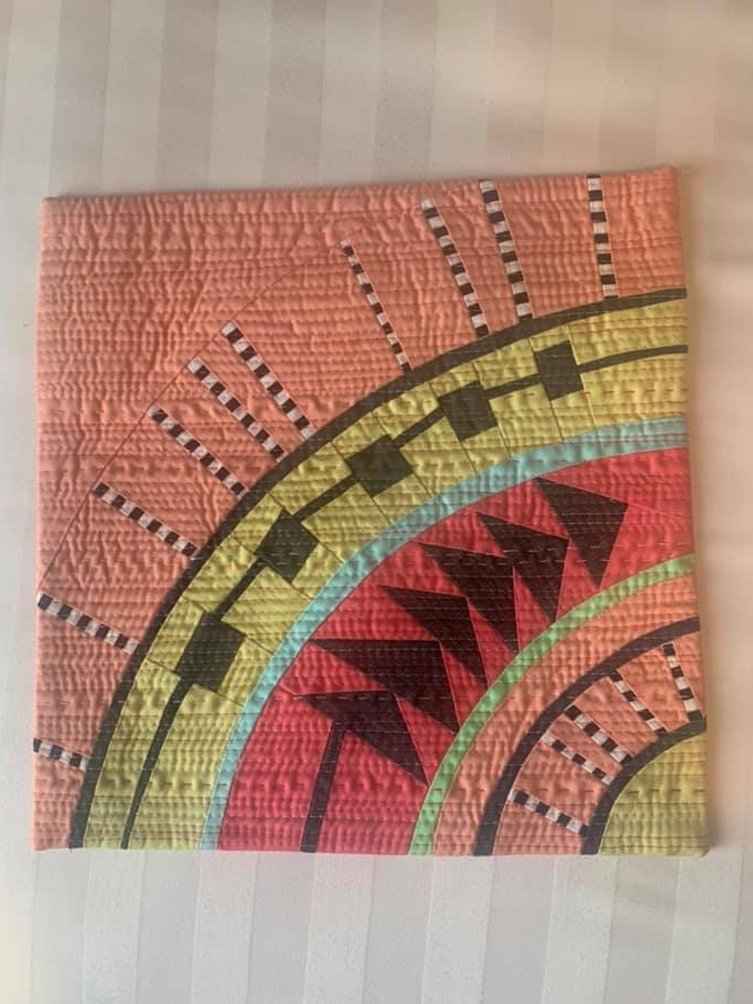 A small, square quilt with a vibrant, abstract design. It features a coral-coloured background with a circular pattern in the center. The circle is filled with various shapes and lines in black, white, green, and blue. The background has a textured, quilted pattern.
