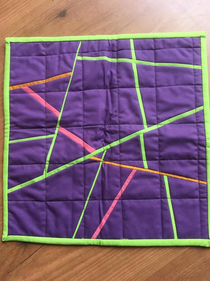 A small, square quilt with a purple background and a green border. The quilt features a geometric design with intersecting lines in pink, orange, and green. The background fabric has a quilted pattern.