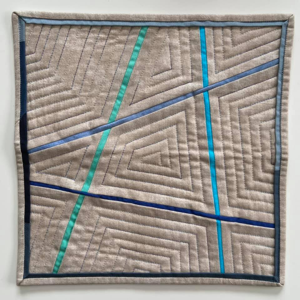 A small, square quilt with a light grey background and a blue border. The quilt features a geometric design with intersecting lines of blue, teal, and dark blue fabric. The lines create triangles and other shapes within the quilt.