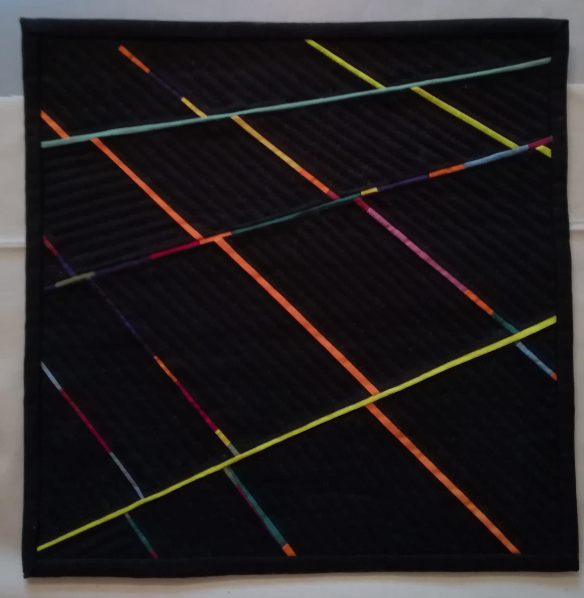 A small, square quilt with a dark background and a vibrant, geometric design. The design consists of colourful, diagonal lines in various shades of orange, yellow, green, and purple. The lines intersect and create a dynamic pattern.