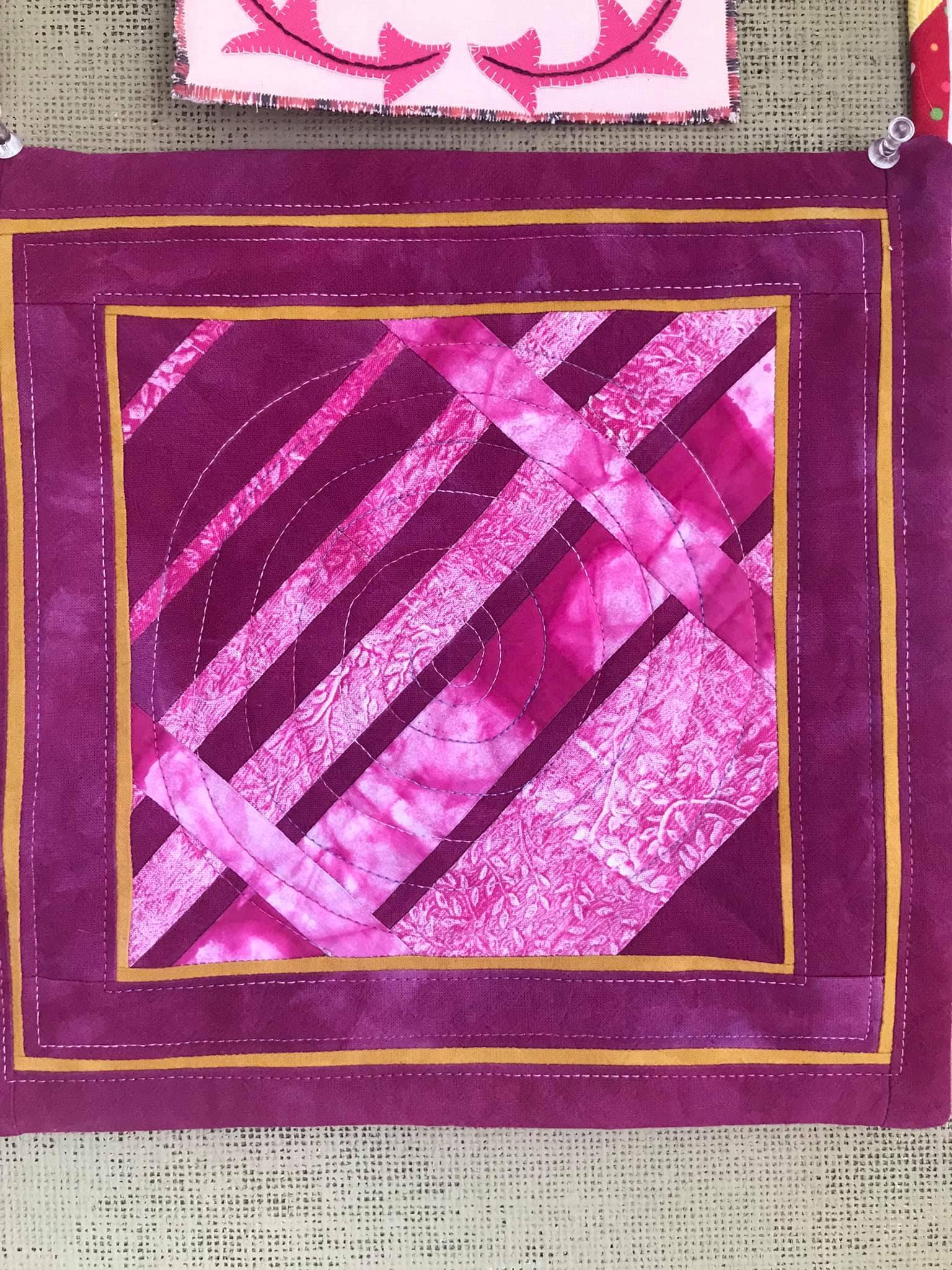 A small, rectangular quilt with a vibrant pink background and a gold border. The quilt features a geometric design with intersecting lines and a circular pattern in the centre. The fabric has a textured, tie-dyed appearance.