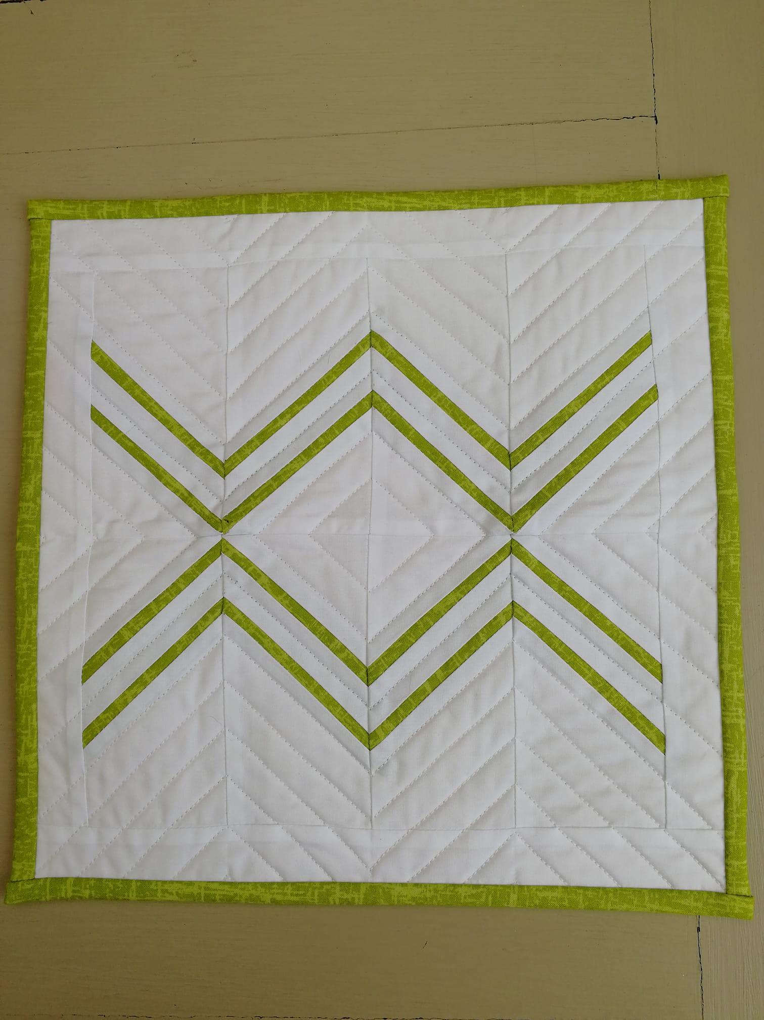A small, rectangular quilt with a white background and a lime green border. The quilt has a geometric design with three sets of lime green lines arranged in a diamond pattern. The lines are quilted in a straight line pattern.