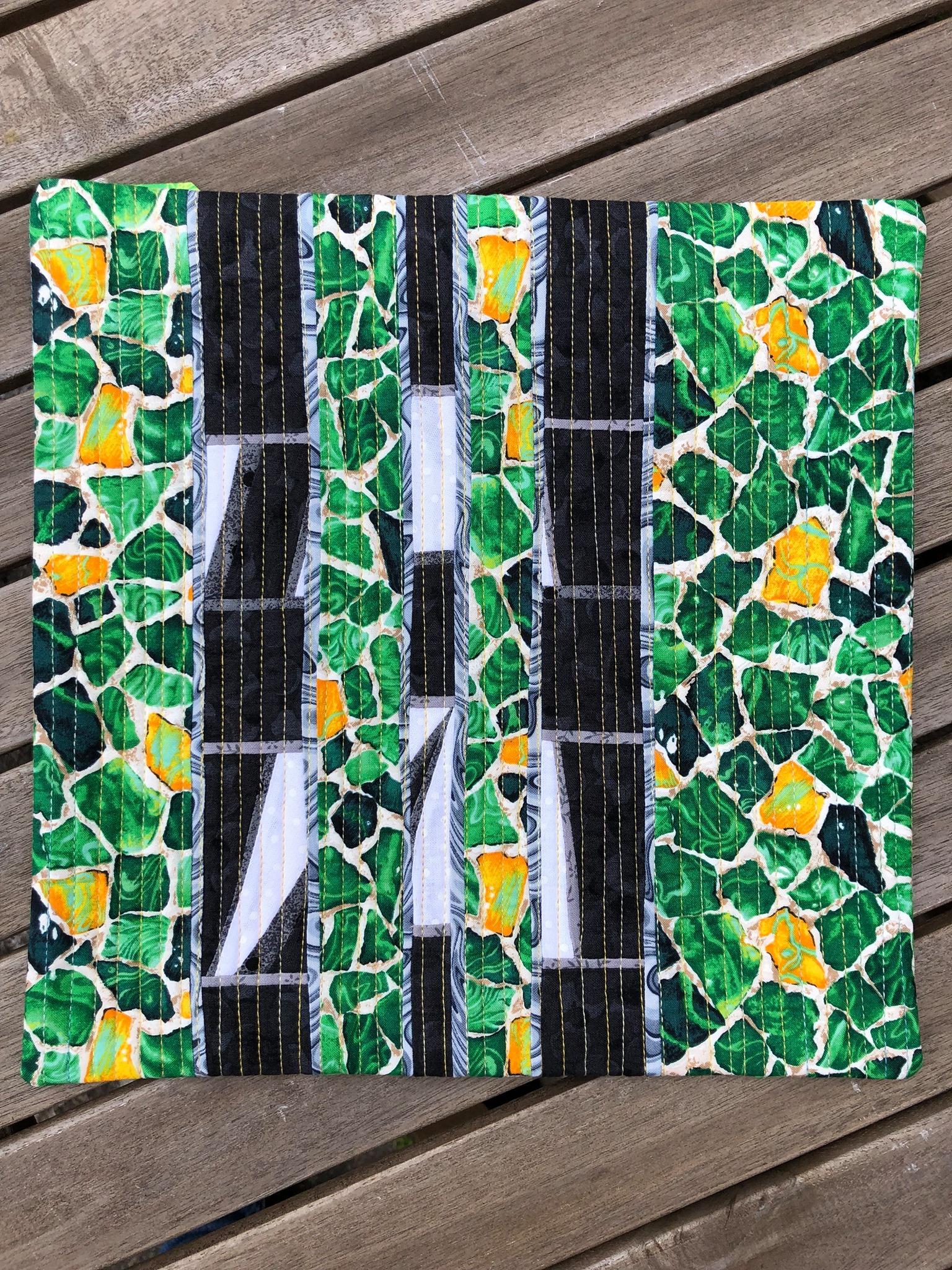 A small, rectangular quilt with a vibrant, abstract design. It features a black background with sections of green and yellow fabric. The green and yellow sections have a pattern of small, irregular shapes.