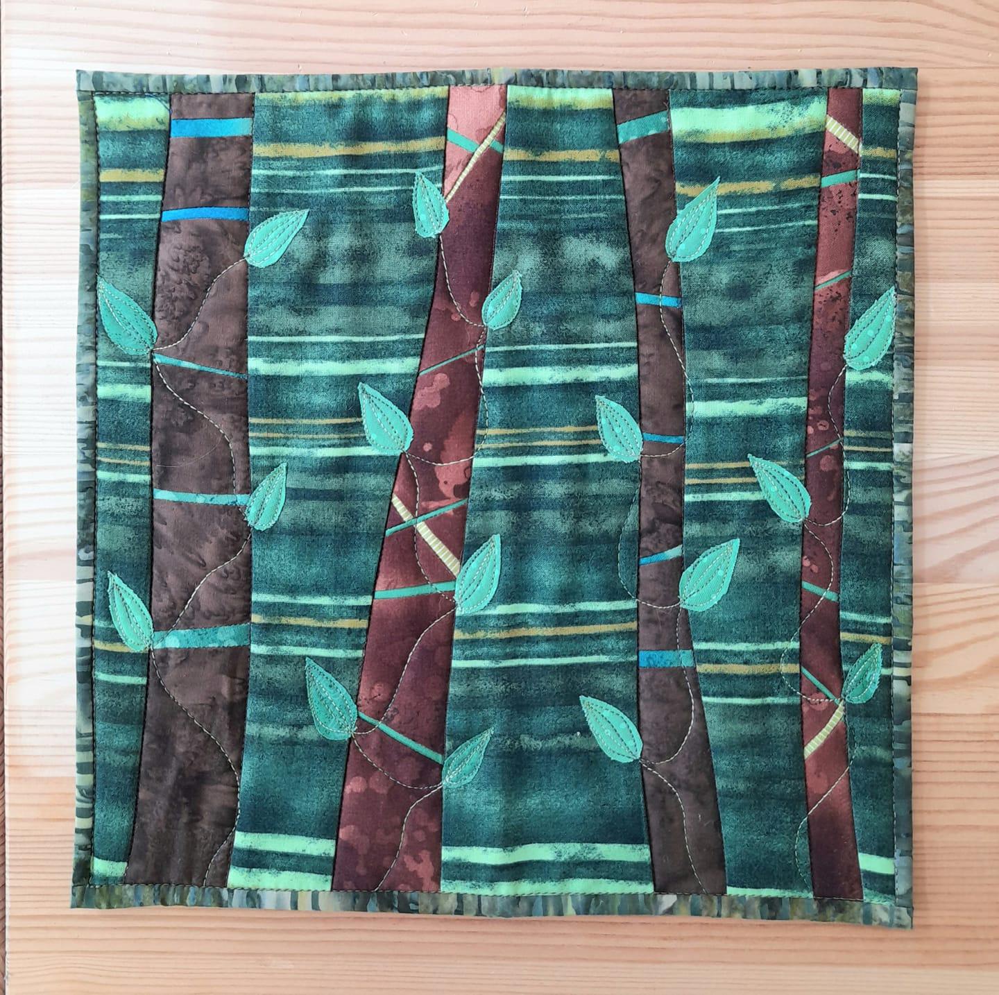 A small, rectangular quilt with a nature-inspired design. It depicts a forest scene with several trees, each with a brown trunk and green leaves. The background is a dark green with lighter green and brown stripes.