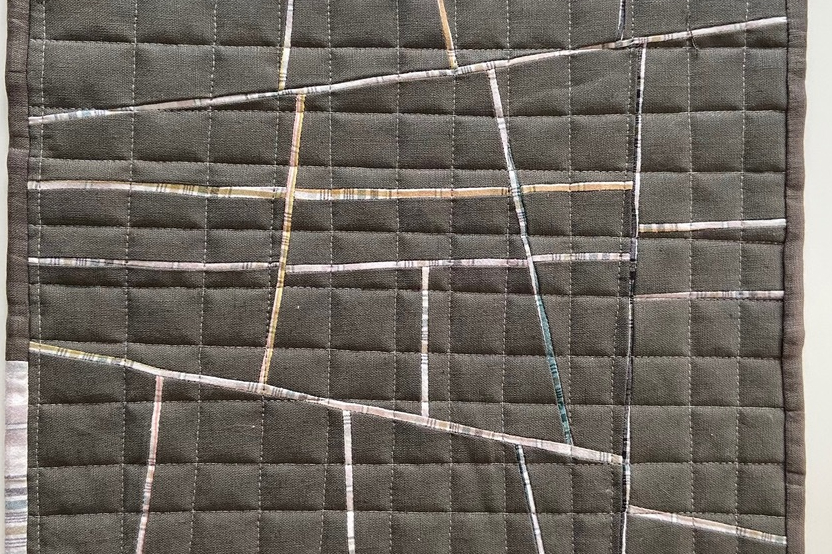 A small, square quilt with a patchwork design. The quilt is primarily dark grey with thin, intersecting lines of light grey, white, and a few pops of tan. The bottom right corner has a small strip of a light plaid fabric.