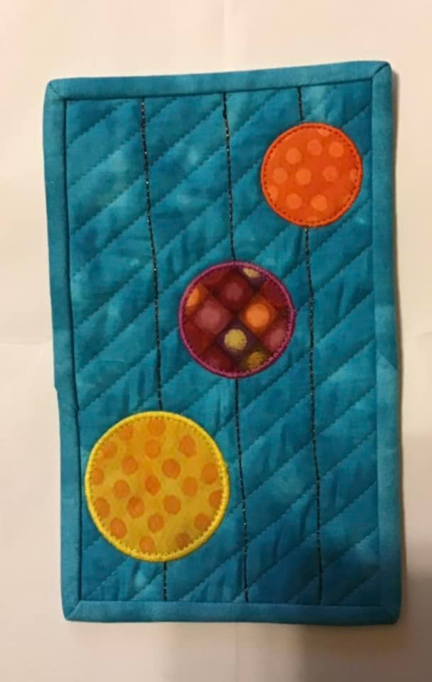 A rectangular quilt with a blue background. Three colorful circles, in orange, red, and yellow, are attached to the background with black thread, resembling ornaments. The background is quilted with a diagonal line pattern.