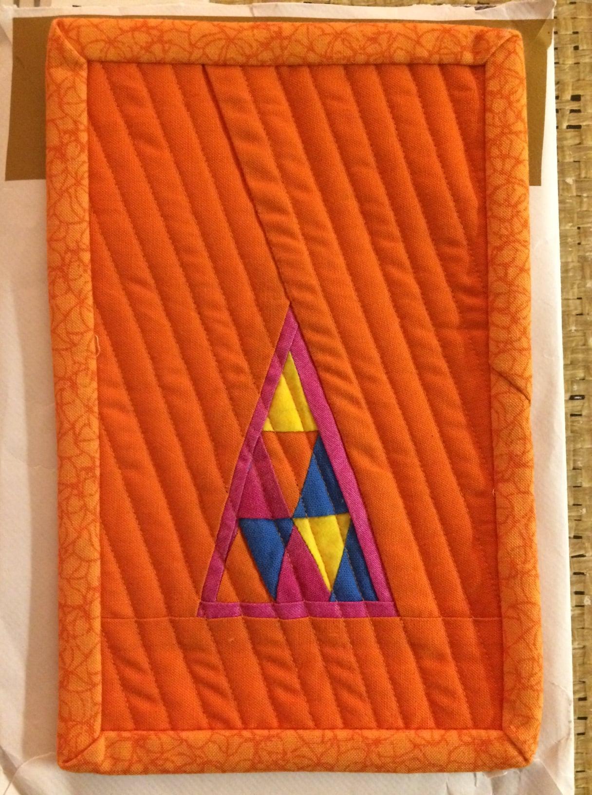 A rectangular quilt with an orange background and a brown border. The quilt features a triangle made up of smaller triangles in pink, yellow, and blue. The background is quilted with diagonal lines.