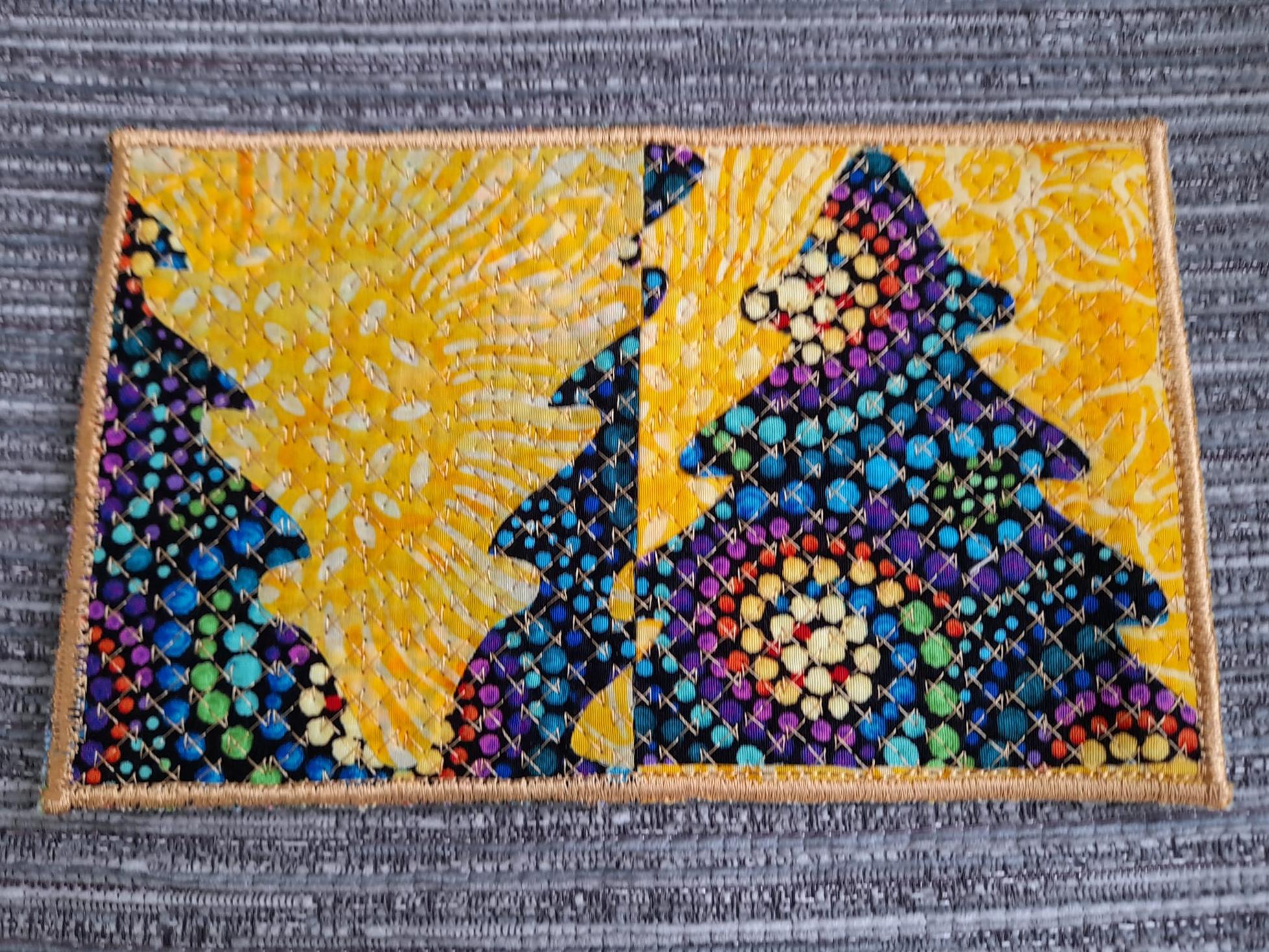 A rectangular quilt with a gold border. The quilt is divided into two sections. The left side features a dark blue tree silhouette against a bright yellow background with a sunburst pattern. The right side shows a colourful tree made up of small, multicoloured shapes.