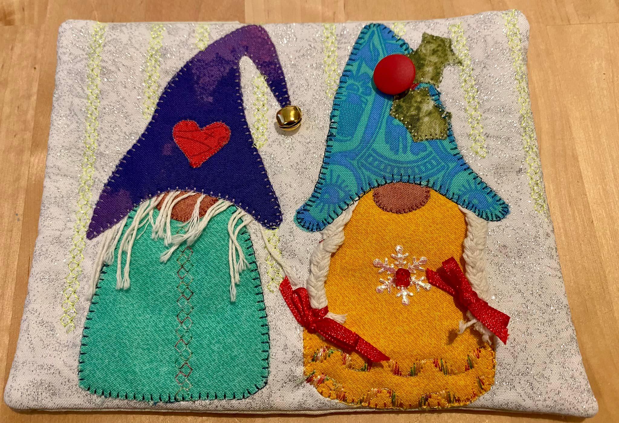 A square quilt with a white background. The quilt features two gnomes standing side by side. The gnome on the left has a purple hat and a green outfit, while the gnome on the right has a blue hat and a yellow outfit. Both gnomes have white beards and red accents. The background is quilted with a subtle, sparkly pattern.