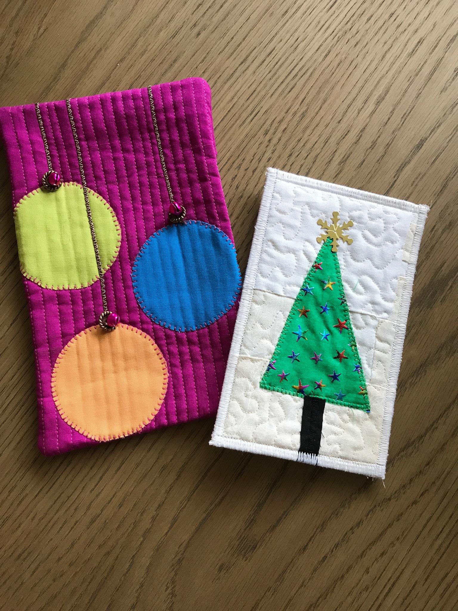 A pink fabric pouch with three colourful circles attached by gold chains, resembling Christmas ornaments and a rectangular quilt with a white background featuring a green Christmas tree with a gold star on top. The tree is decorated with small, colourful shapes.
