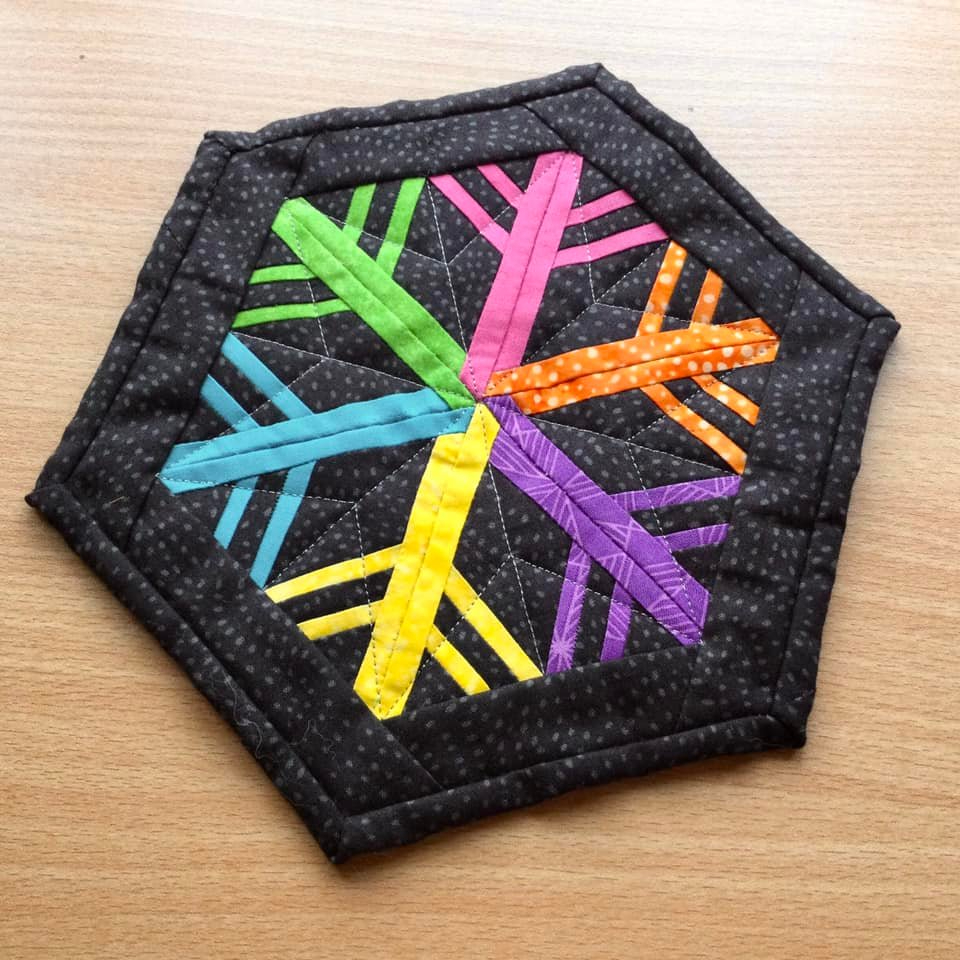 A hexagonal quilt with a black background. The quilt features a colorful snowflake design with arms in green, pink, blue, orange, and purple. The background is quilted with a subtle, dotted pattern.