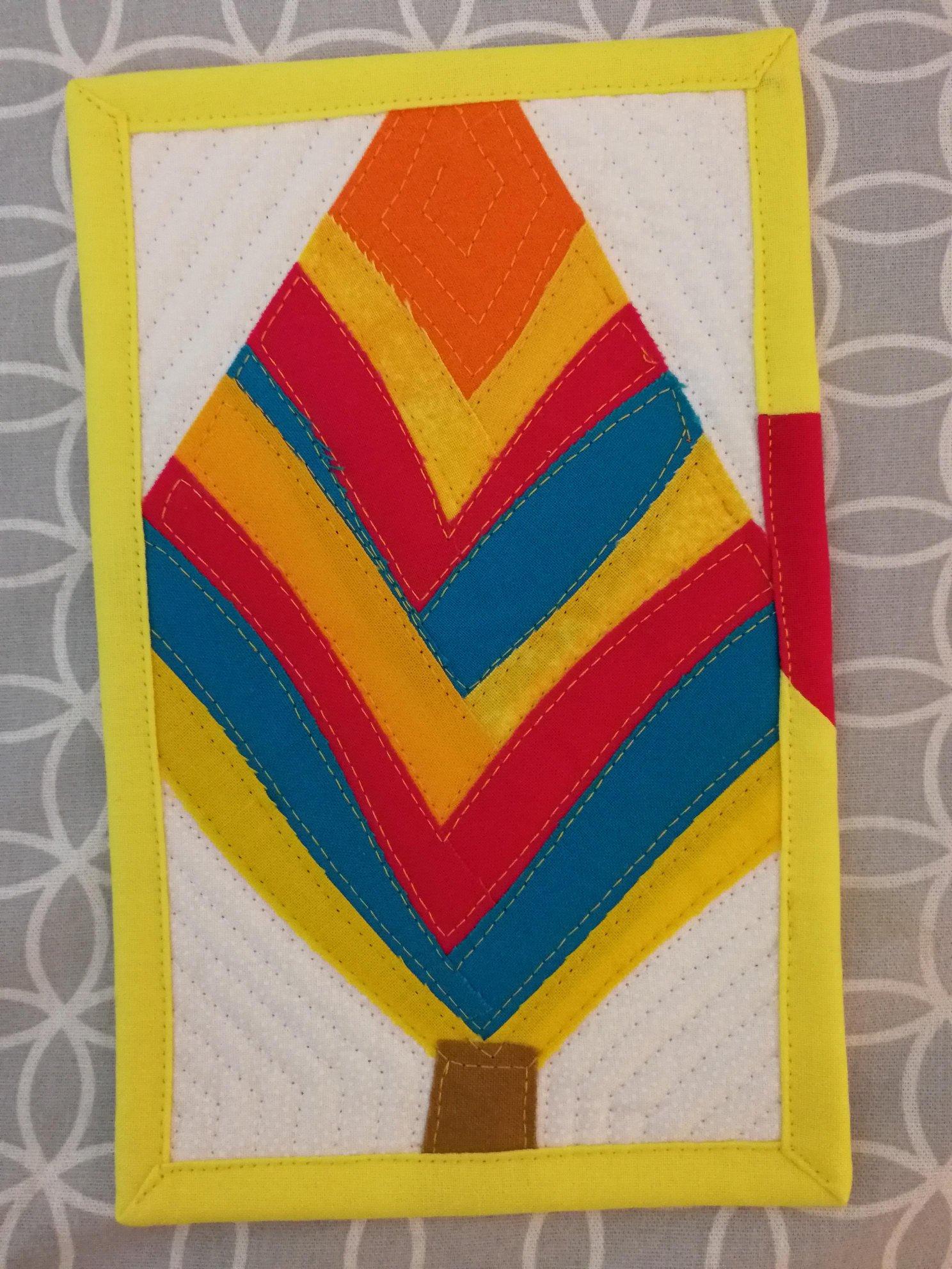 A square quilt with a yellow border. The quilt features a colorful, abstract tree design with layers of red, orange, yellow, and blue fabric. The tree trunk is brown. The background is a white fabric with a subtle texture.