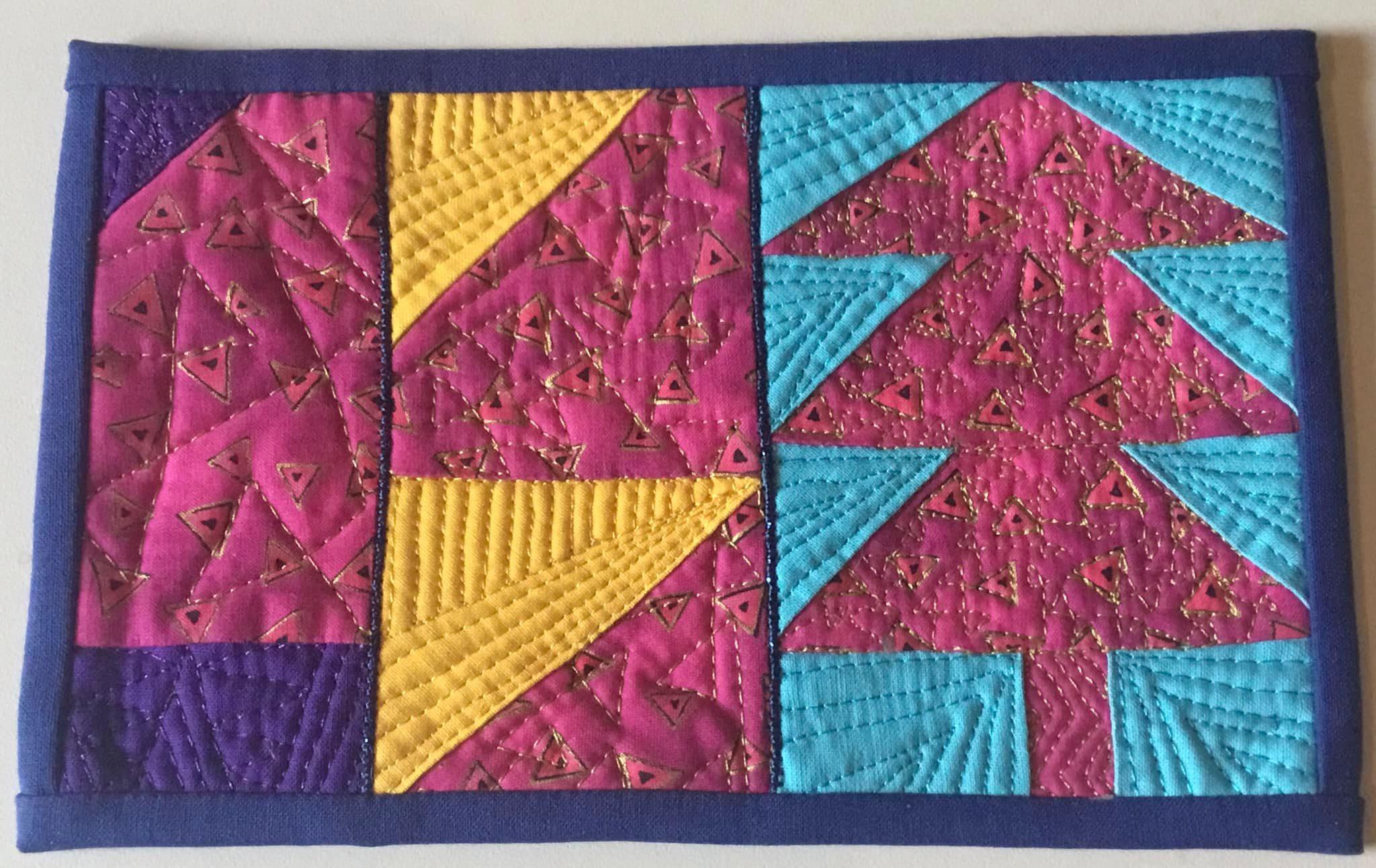 A rectangular quilt with a blue border. The quilt features a patchwork design with triangles in shades of pink, yellow, and blue. The triangles are arranged in different patterns, creating a visually interesting composition. The background is quilted with a variety of lines and shapes.