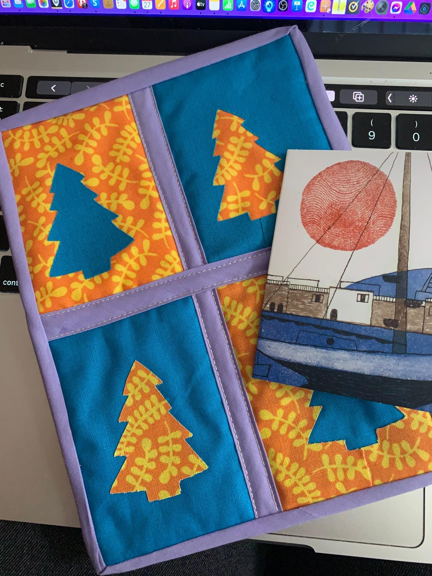 A square quilt with a patchwork design. The quilt is divided into four sections, each with a different color background: orange, blue, orange, and blue. On each background, there is a green Christmas tree shape. Next to the quilt is a card with a drawing of a boat and a red sun.