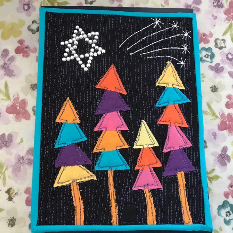 A square quilt with a black background and a turquoise border. The quilt features a night sky scene with a silver star and shooting stars, and a row of colorful, striped trees.
