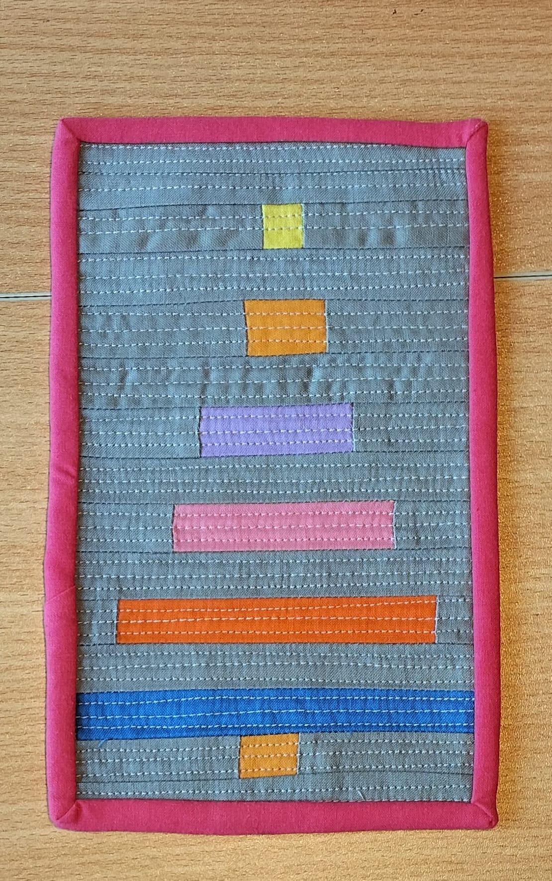 A square quilt with a red border. The quilt features a colorful, abstract design with horizontal strips of fabric in various colors, including yellow, orange, purple, pink, and red. The background is a textured grey fabric.