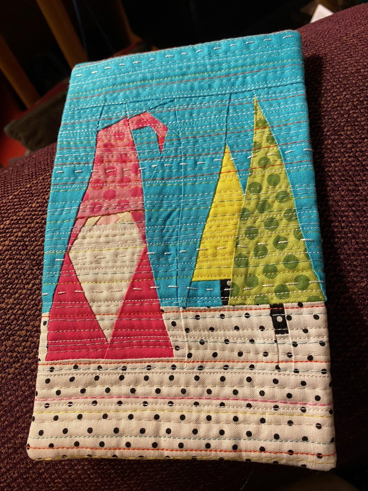 A small, rectangular quilt with a festive winter scene. The background is a bright blue, representing the sky. A pink gnome with a white beard stands on a snowy ground, which is depicted with a black and white polka dot pattern. Behind the gnome are two Christmas trees, one yellow and one green.