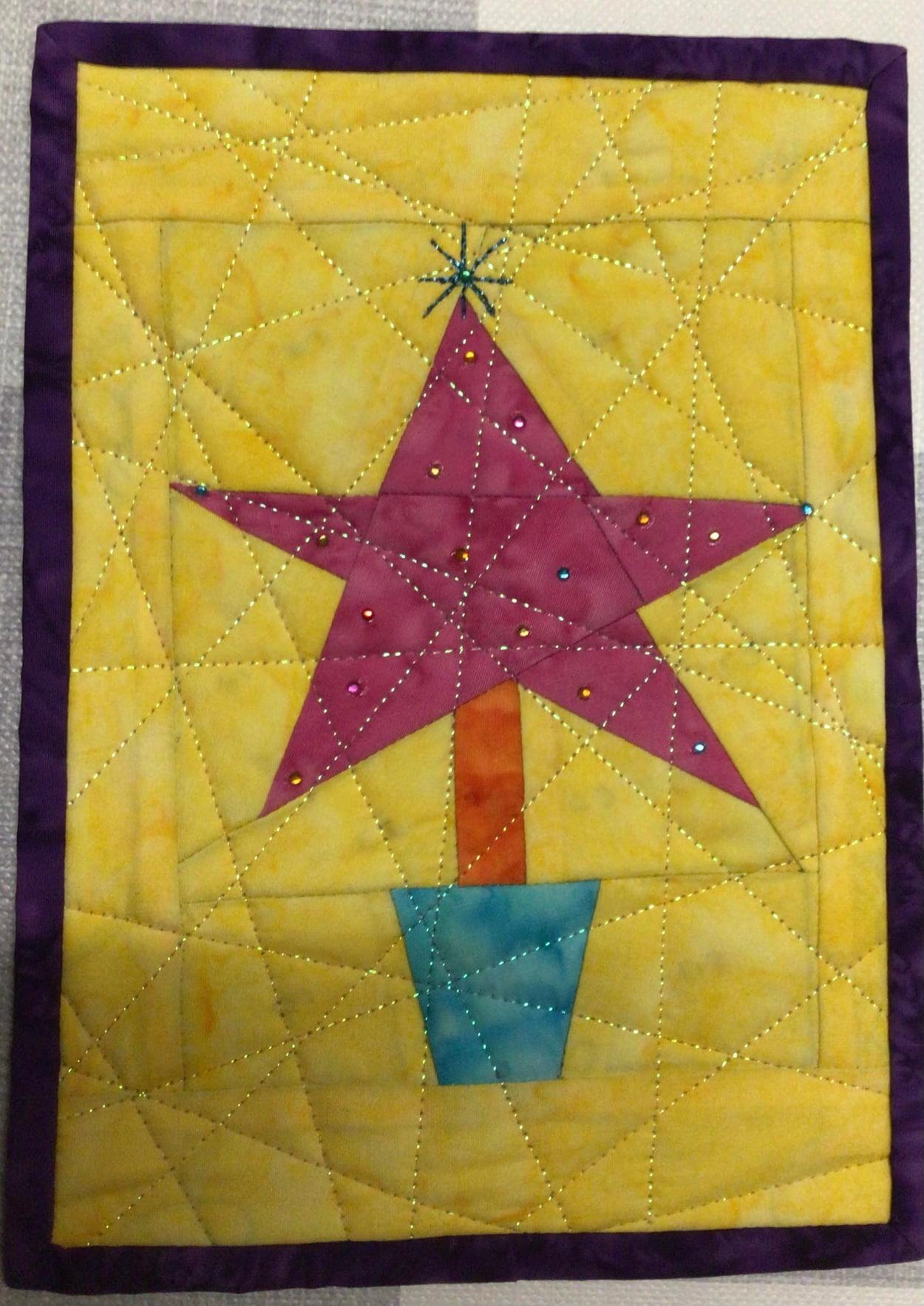 A square quilt with a purple border. The quilt features a pink star-shaped tree on a yellow background. The tree has a blue pot and a silver star on top. The background is quilted with a web-like pattern.