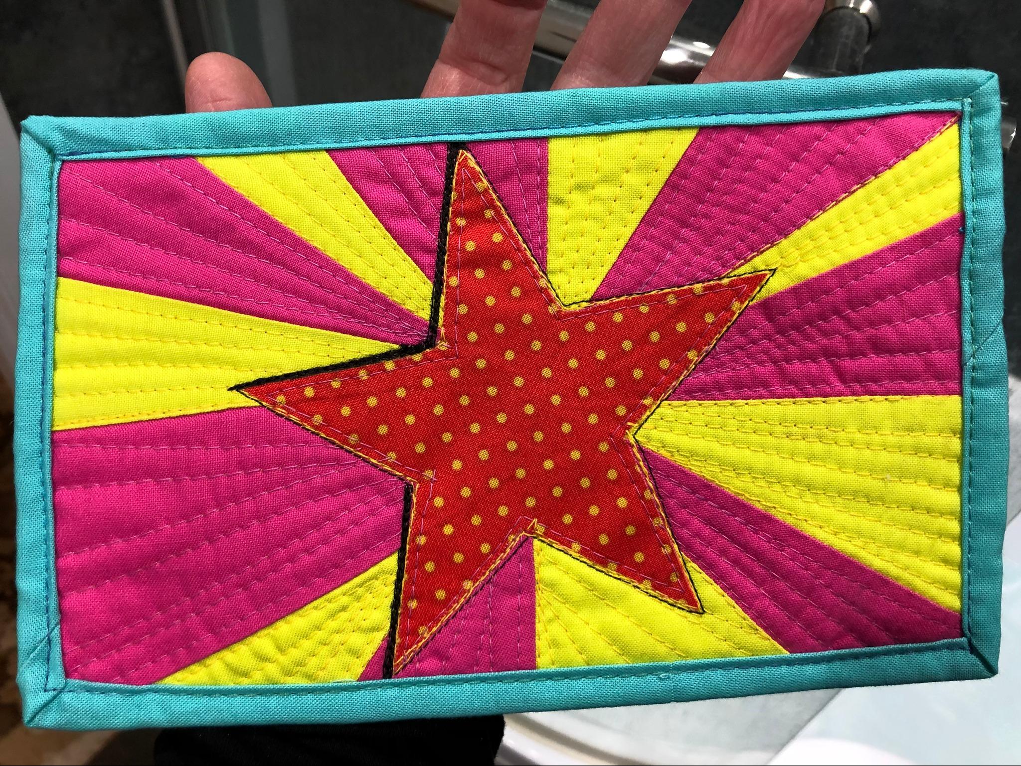 A rectangular quilt with a turquoise border. The quilt features a large, red, polka-dot star in the center, surrounded by alternating pink and yellow stripes radiating outwards. The background is quilted with straight lines.