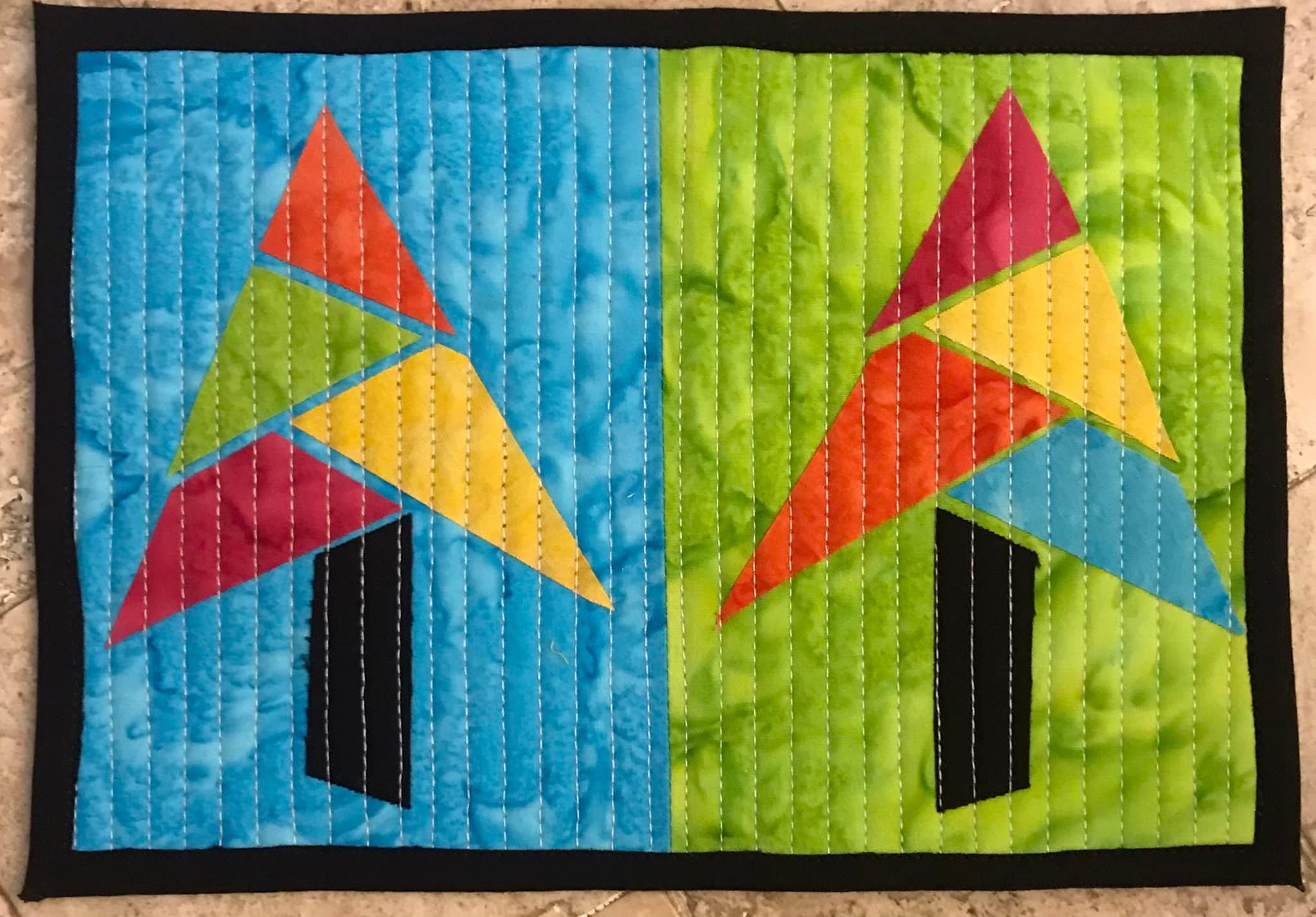 A rectangular quilt with a black border. The quilt is divided into two sections, each with a different colored background: blue on the left and green on the right. On each background, there is a colorful, abstract tree made up of triangles in various colors. The background is quilted with vertical lines.