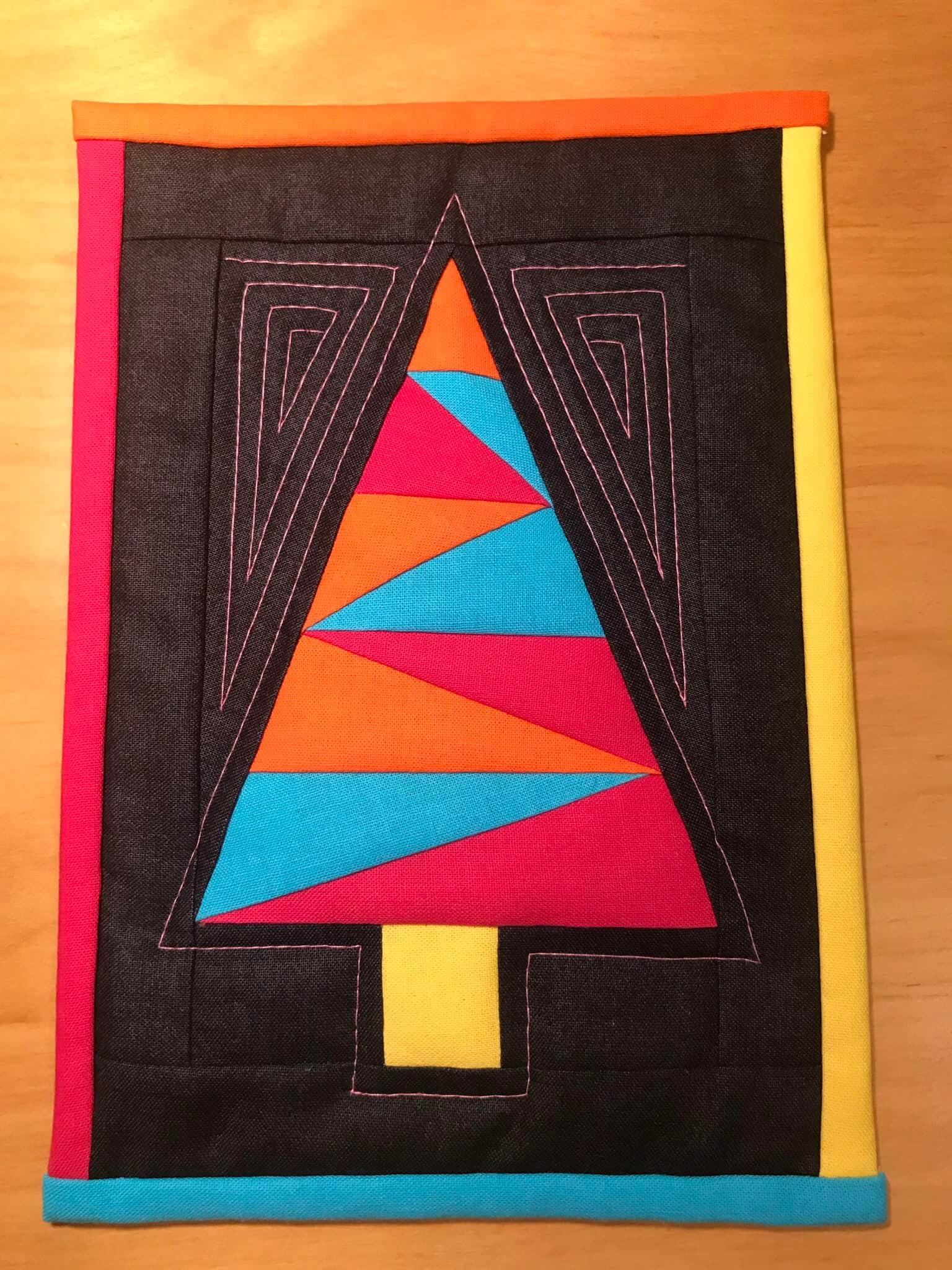 A square quilt with a colorful border. The quilt features a stylized Christmas tree made up of triangles in various colors like red, orange, yellow, and blue. The tree is surrounded by a black frame with a swirling, linear pattern.