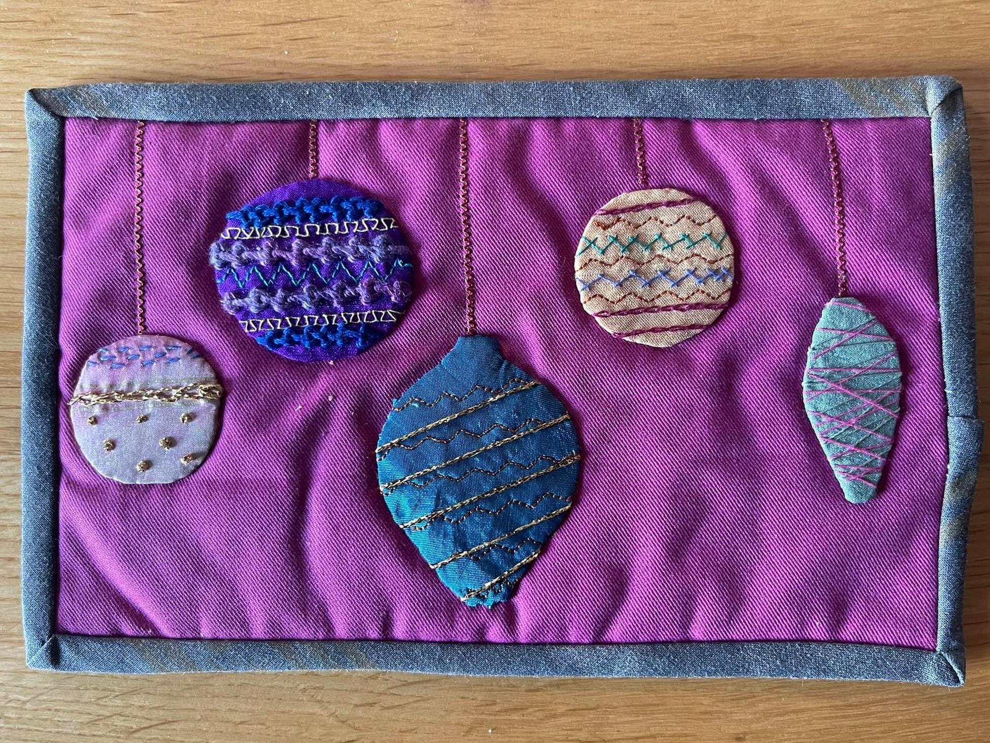 A small, rectangular quilt with a purple background and a blue border. The quilt features four embroidered ornaments hanging from strings. The ornaments are in various shapes and colours, including a circle, an oval, a teardrop, and a diamond.